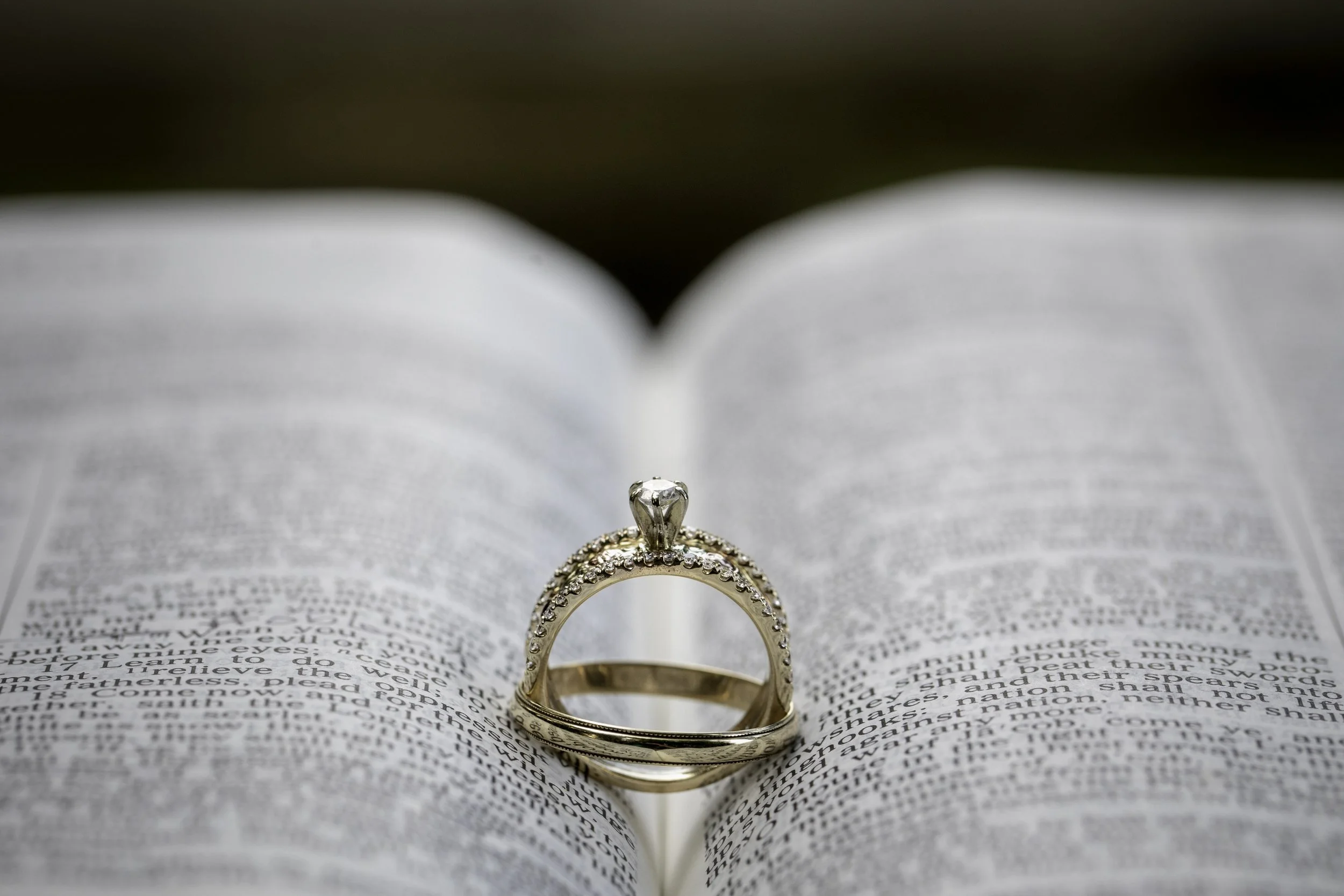 A gold engagement ring with diamonds is placed on the center of an open Bible, creating a heart shape with the pages.