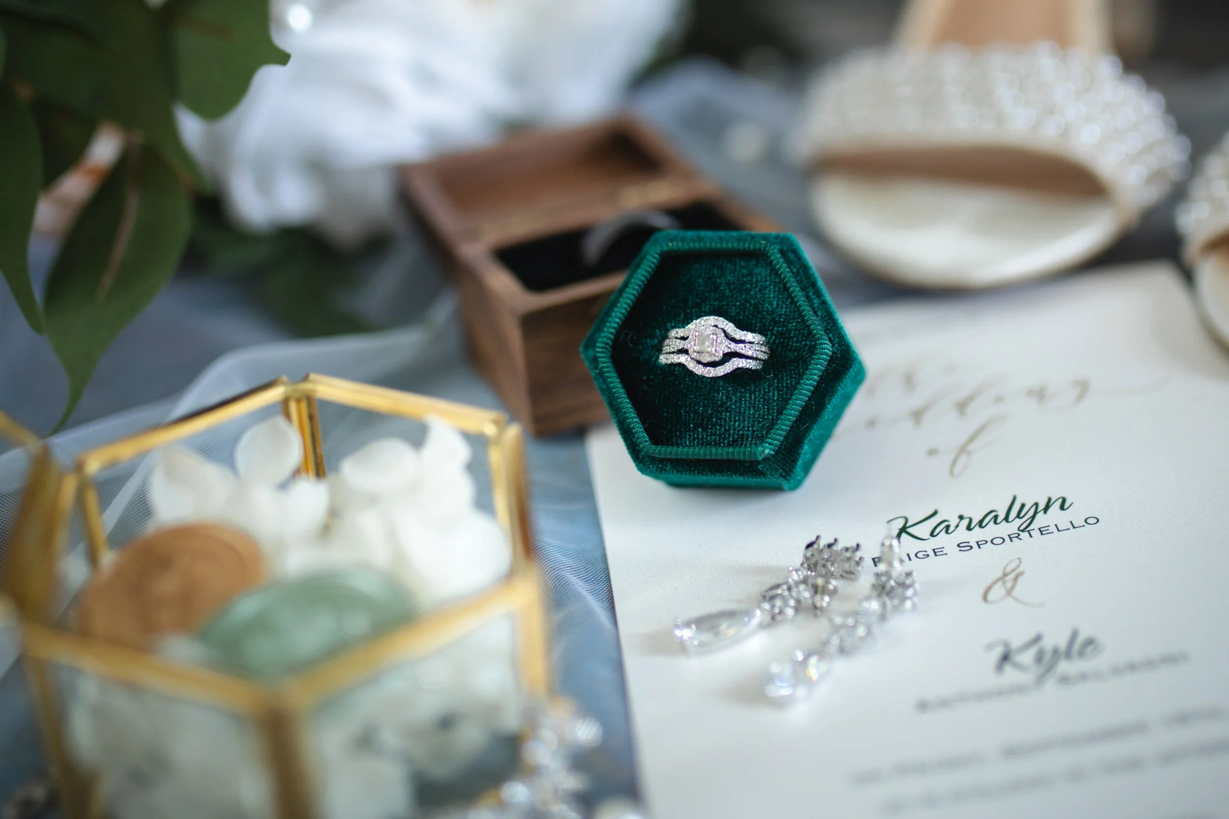 A diamond ring in a green velvet box, a pair of earrings, and a wedding invitation with the names Karalyn and Kyle.