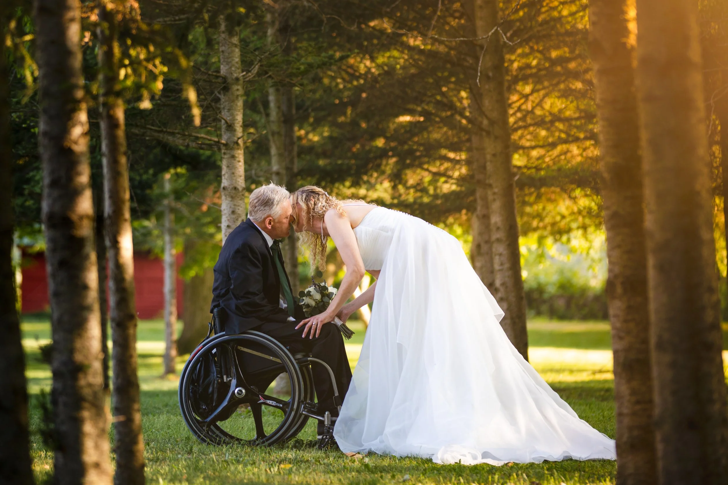 A bride in a white wedding gown leaning over and kissing a man in a wheelchair, who is dressed in a black suit, in a wooded outdoor setting during sunset.