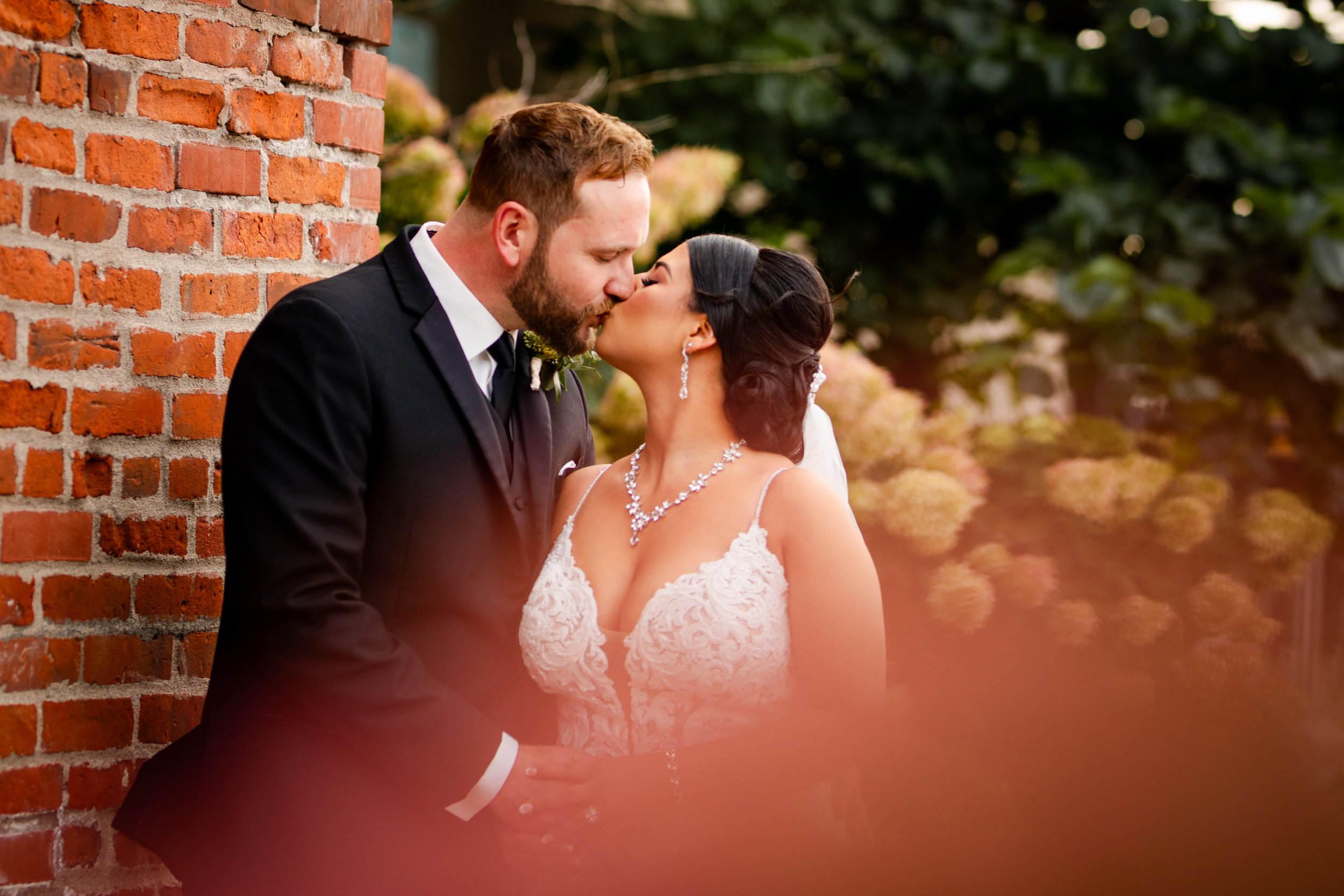 A bride and groom sharing a kiss outdoors, with the groom dressed in a black tuxedo and the bride in a white lace wedding gown and jewelry, standing near a brick wall with greenery and flowers in the background.