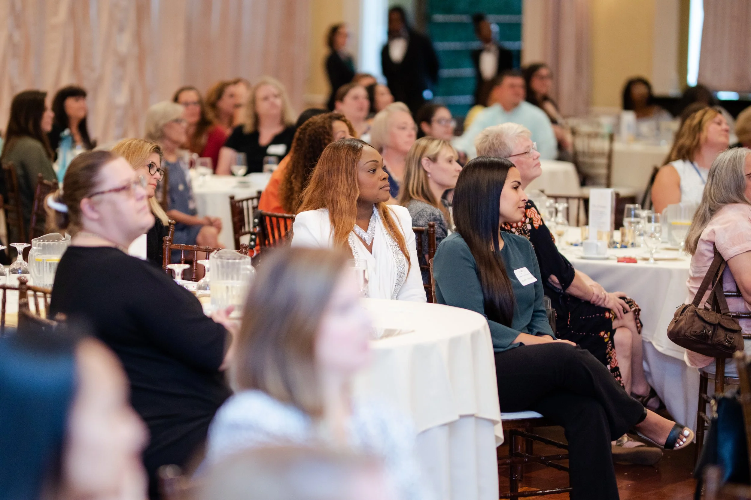 Audience of diverse women seated at round tables, attentively listening during a conference or seminar in a large decorated room.
