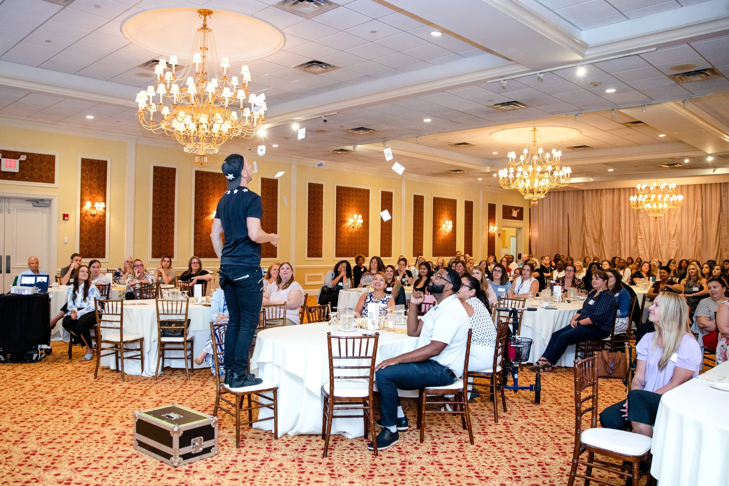 A performer standing on a chair on a stage addressing an audience at a banquet hall with chandeliers and banquet tables.