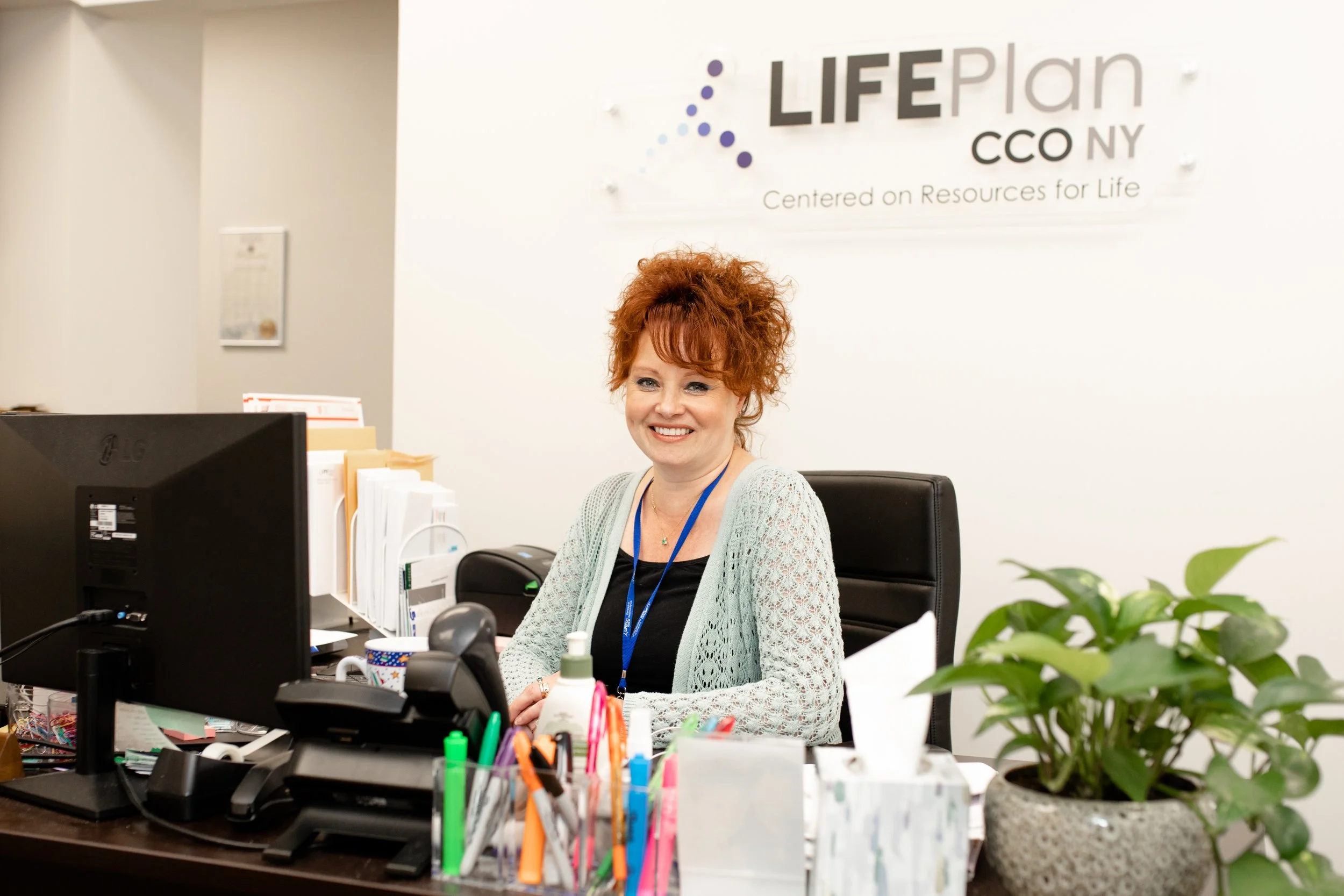 Woman with curly red hair smiling at her office desk, surrounded by office supplies, with a sign on the wall behind her reading 'LIFE Plan CCNY' and the tagline 'Centered on Resources for Life.'
