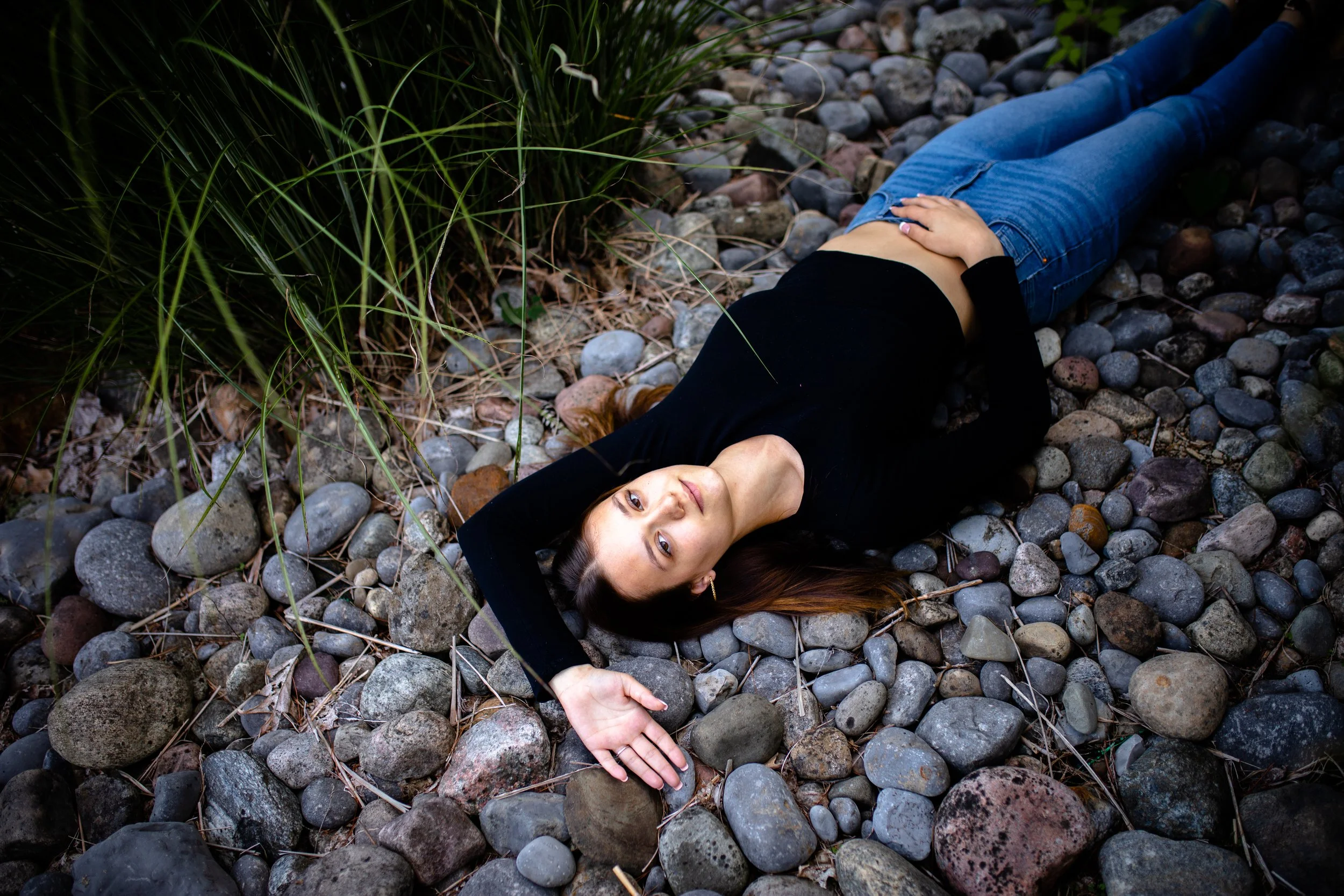 A woman lying on a bed of smooth rocks outdoors, with one arm resting on her forehead and the other on her stomach, wearing a black long-sleeve top and blue jeans, surrounded by green foliage.