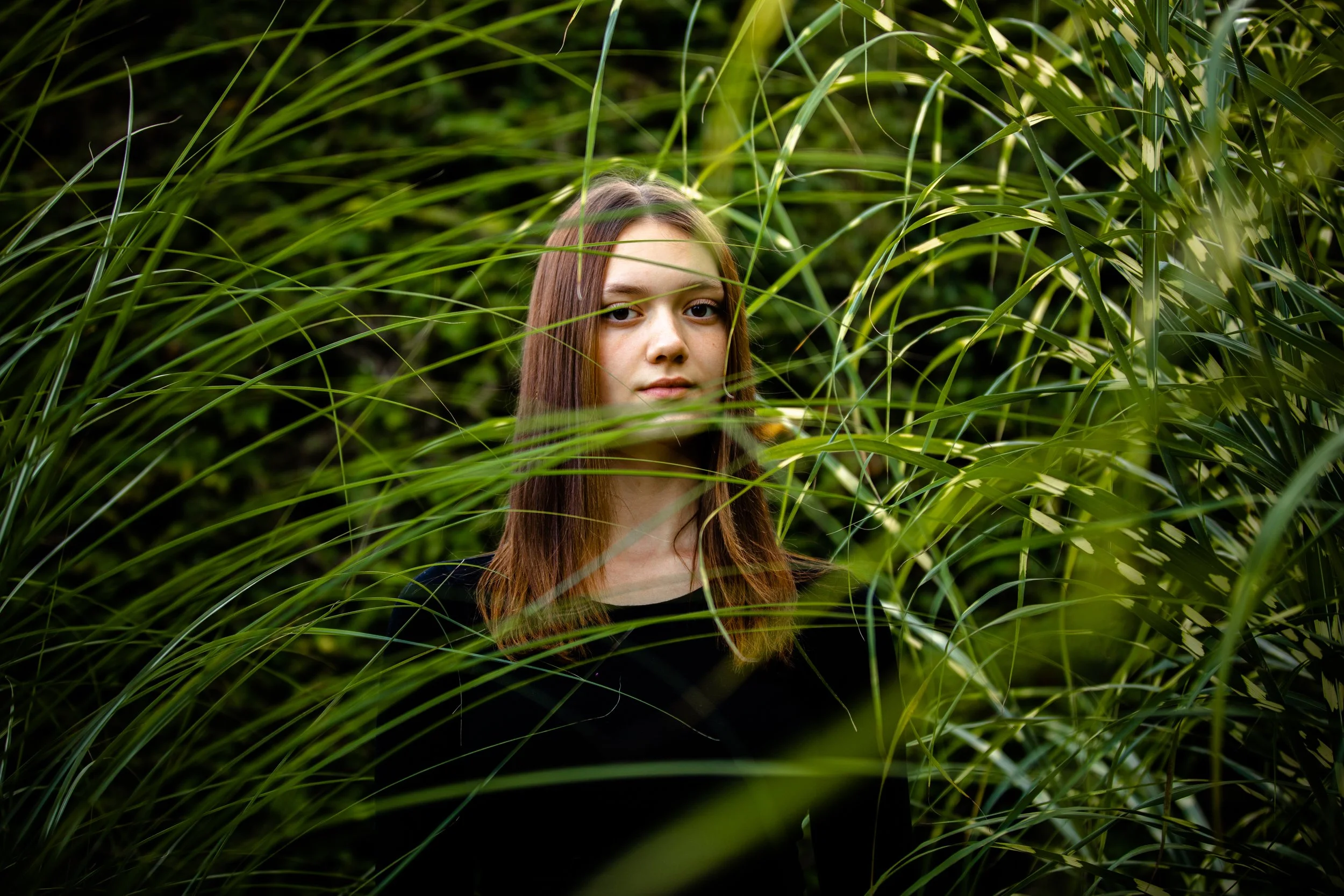 Young woman with long brown hair standing among tall green grass and plants, looking directly at the camera.