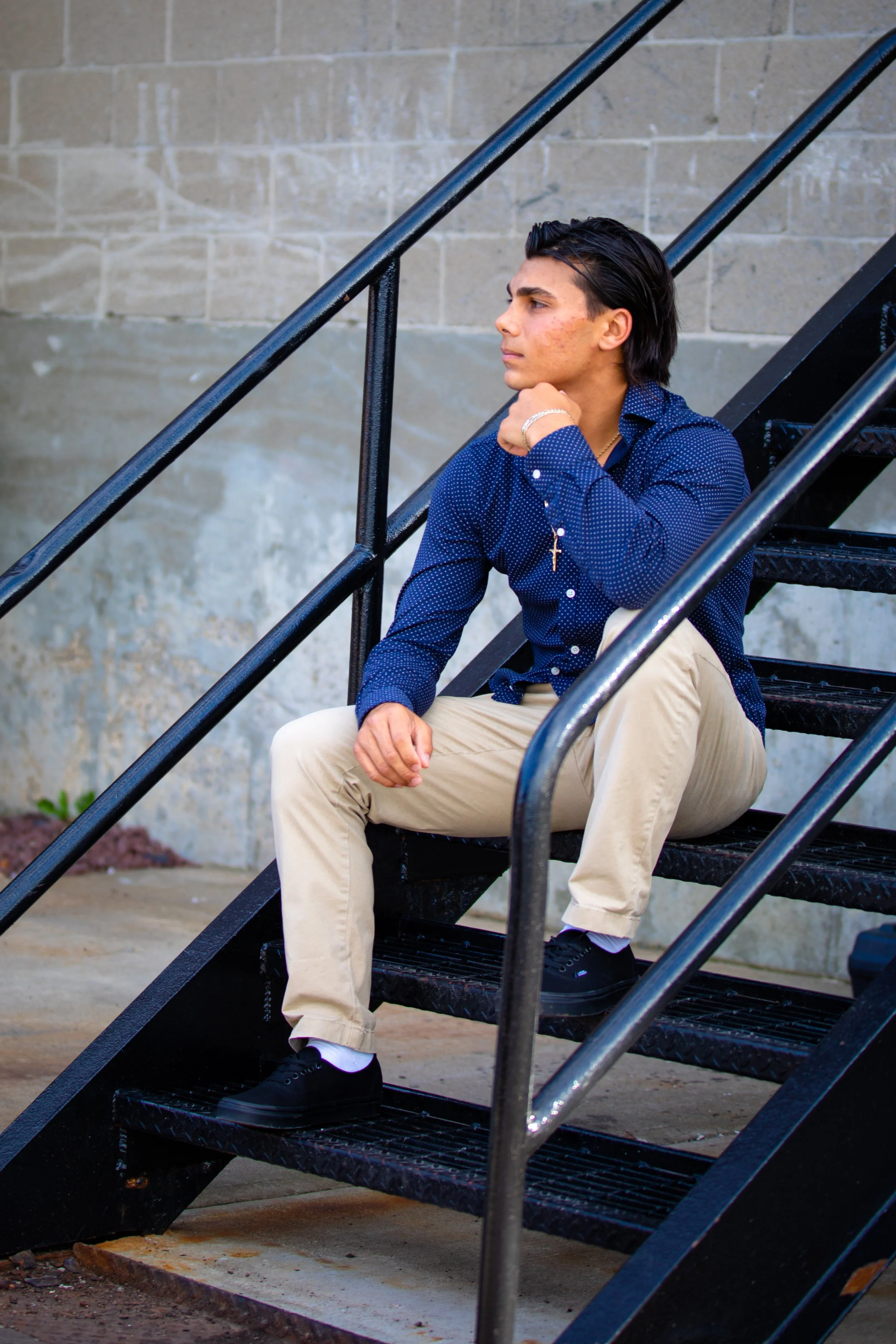 Young man sitting on metal outdoor staircase, resting his chin on his hand, looking thoughtfully to the side, wearing a navy blue shirt, beige pants, black shoes, and a gold cross necklace.