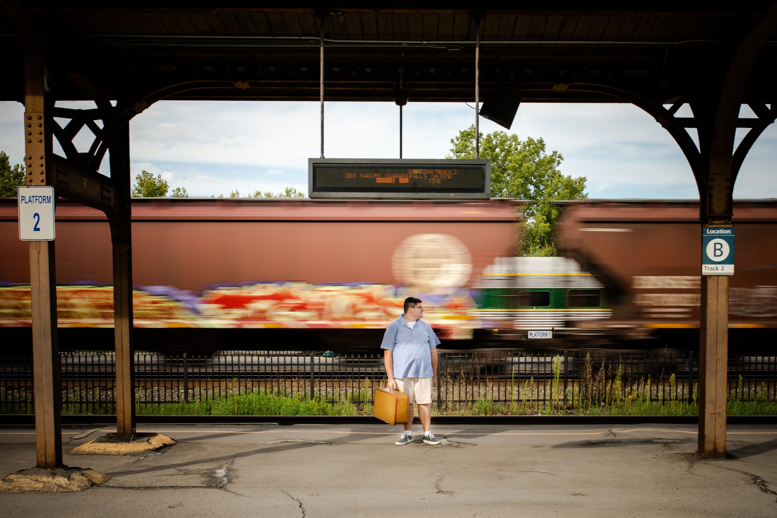 A man holding a suitcase waiting at a train station as a train passes by in the background.