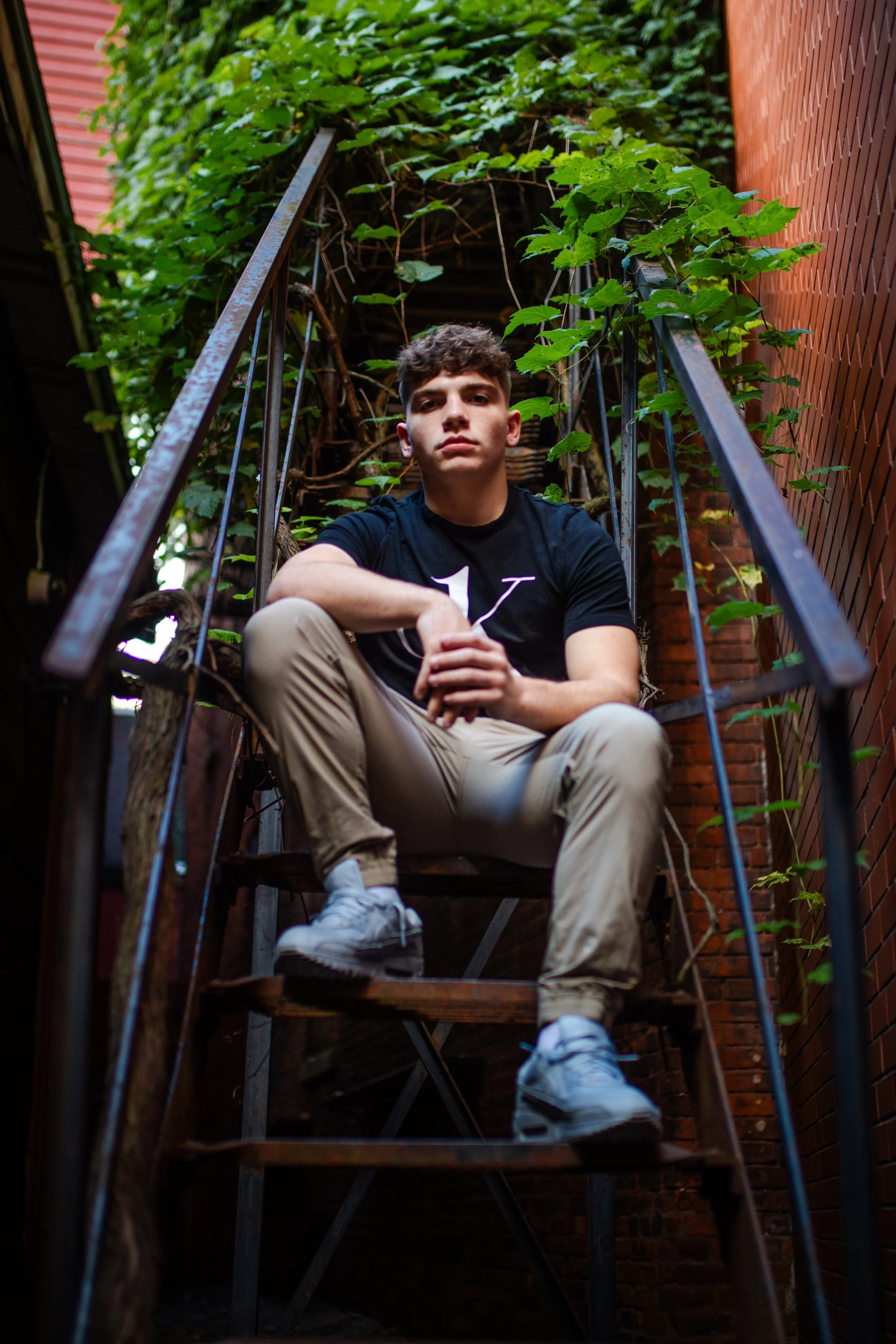 A young man sitting on an outdoor metal staircase with greenery and vines growing around him, between brick walls.