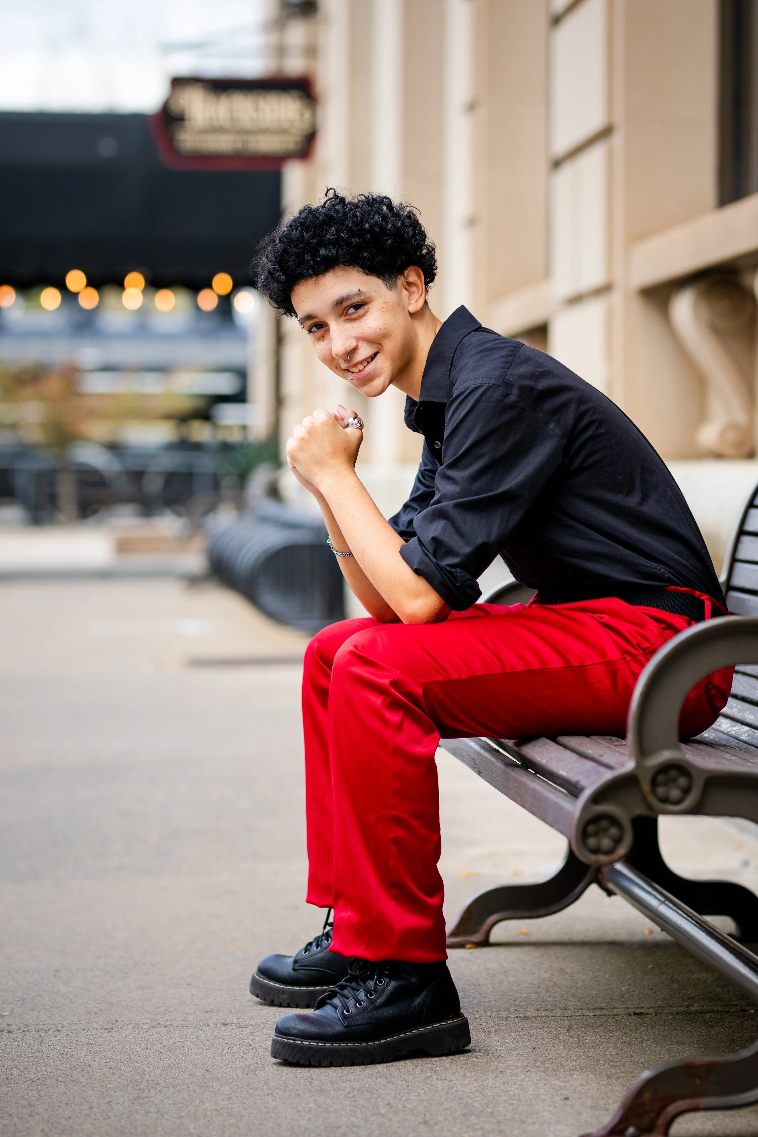 Young person with curly hair sitting on a bench outdoors, smiling, wearing a black shirt and red pants.