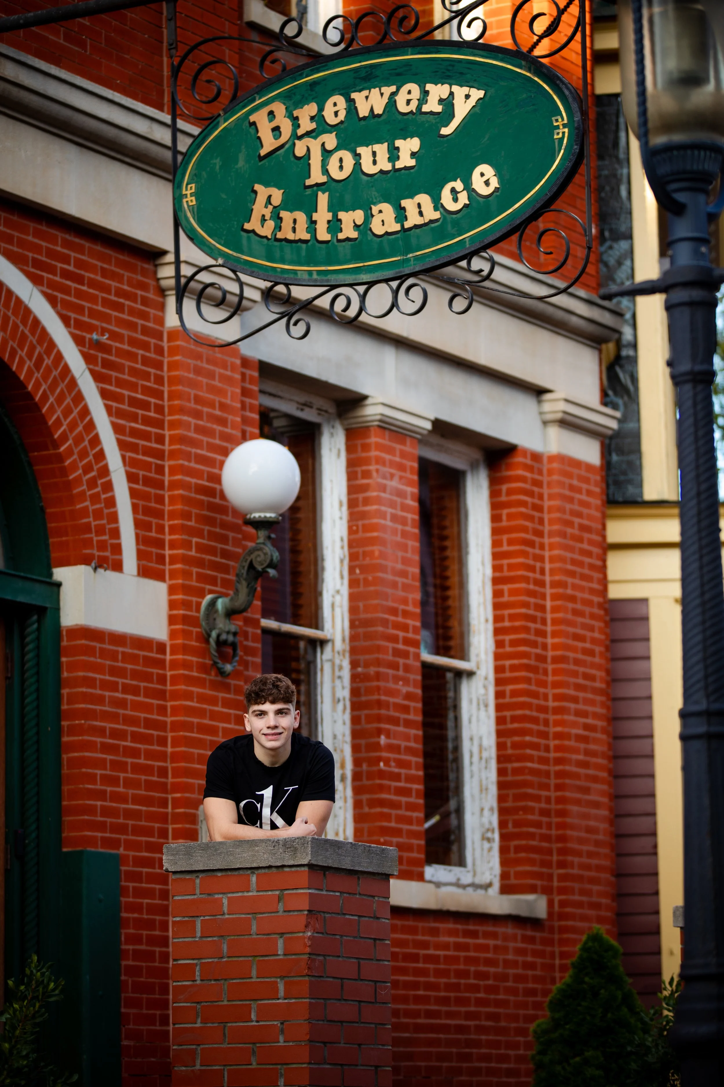 A young man leaning on a brick pillar outside a building with a sign that reads 'Brewery Tour Entrance' in front of a brick building with old wooden window frames and a vintage-style lamp.