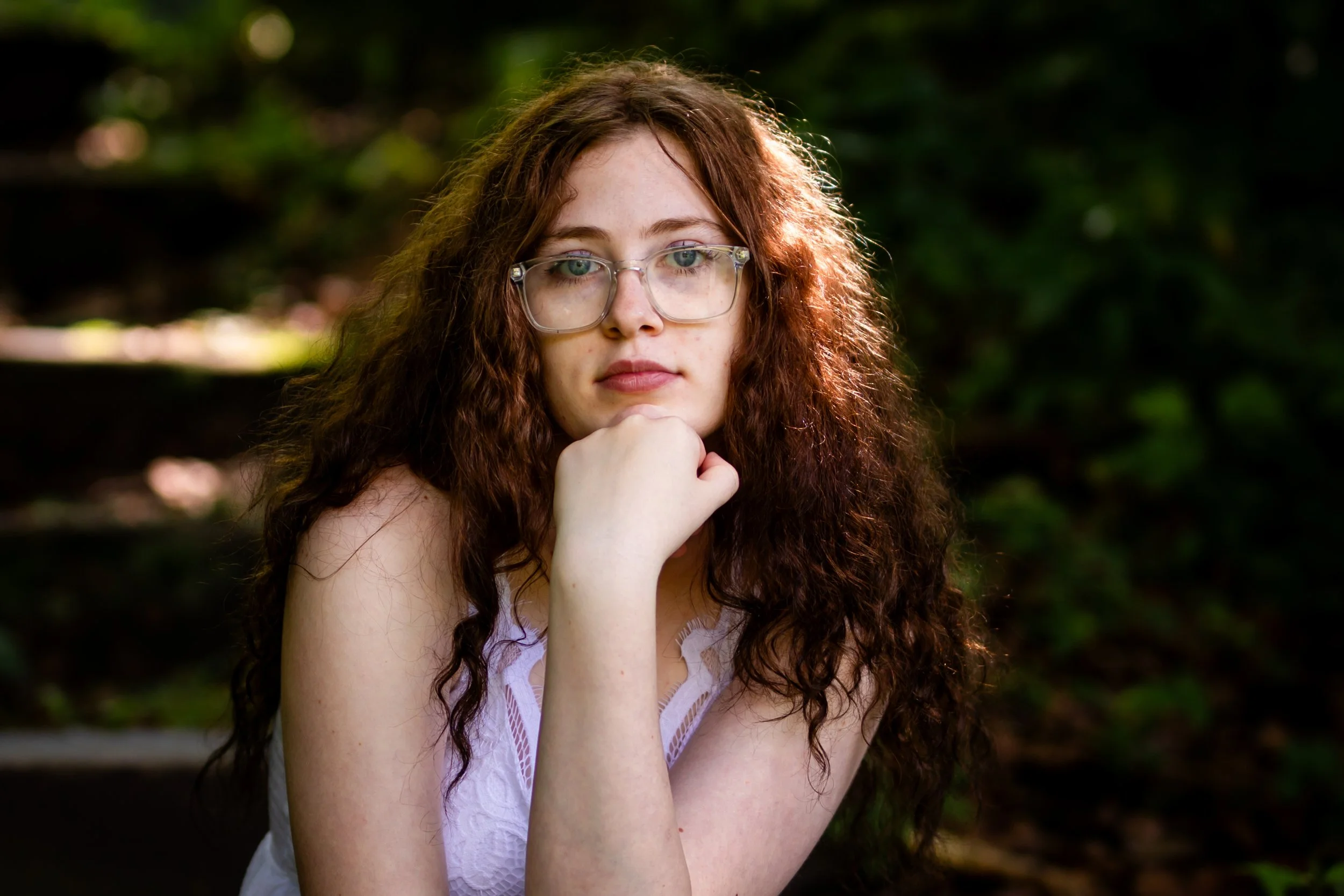 A young woman with curly red hair and glasses posing outdoors with her chin resting on her hand. She is looking directly at the camera, wearing a white lace top, with a blurred natural background.