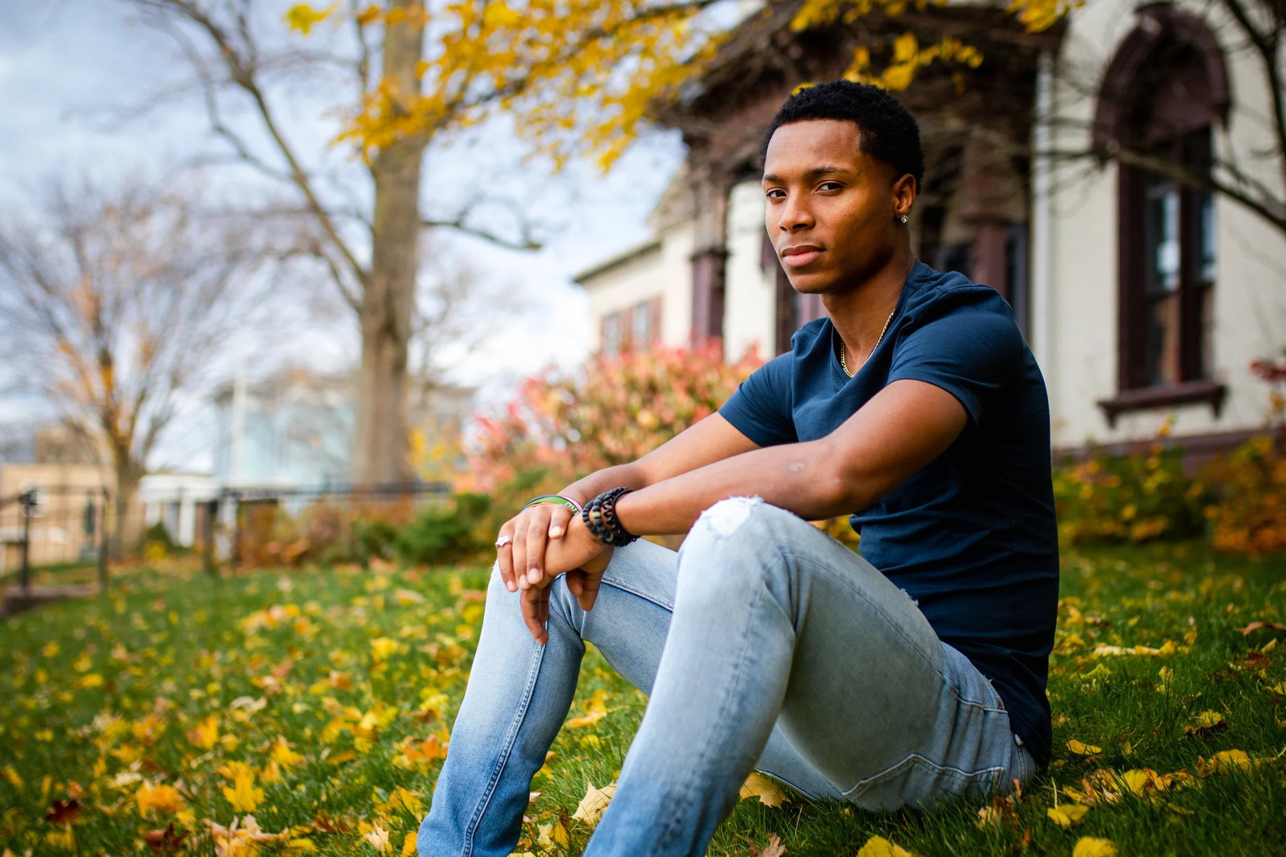 Young man sitting outdoors on autumn grass with fallen leaves, wearing a dark blue T-shirt and ripped jeans, near a house with trees in the background.