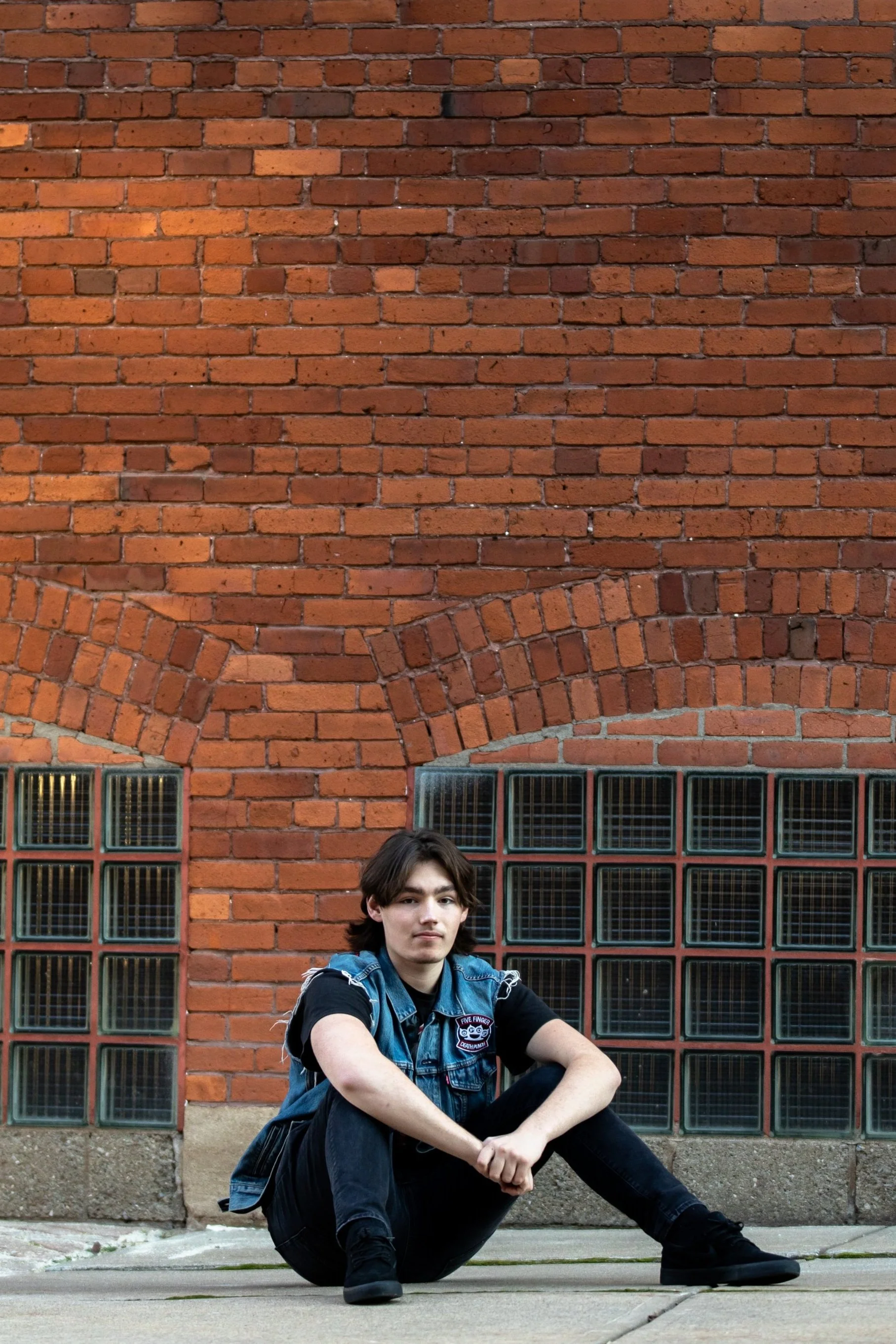 A young man with dark hair wearing a denim vest and black shirt sitting on the sidewalk in front of a brick wall with glass block windows.