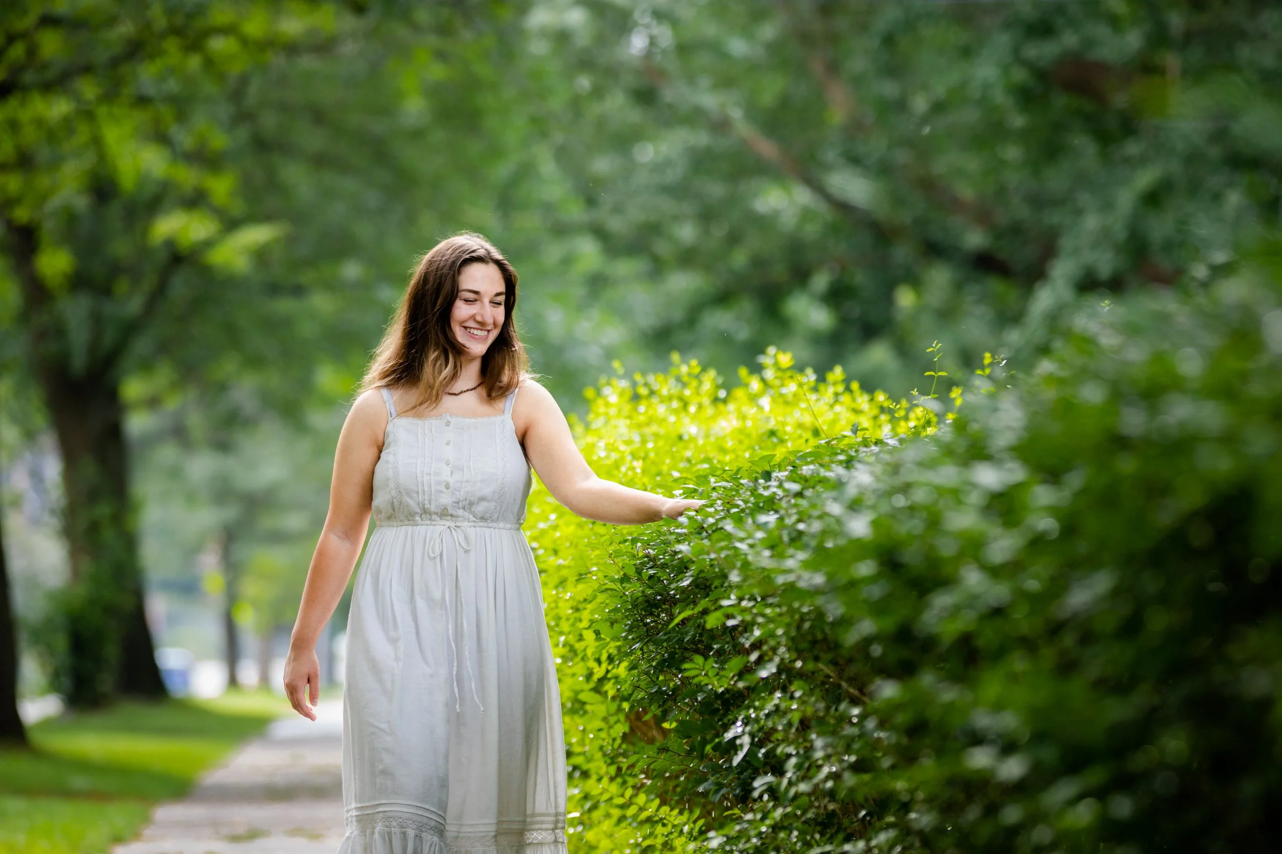 A young woman in a white sleeveless dress smiles as she walks along a sidewalk, touching a green bush on a sunny day.