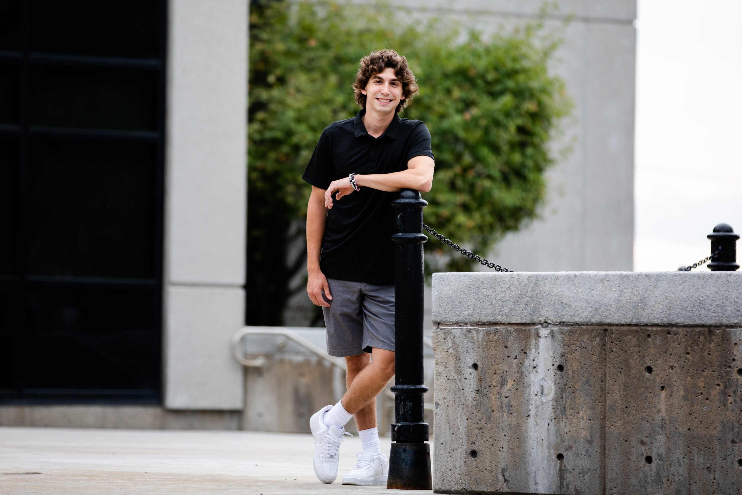 Young man with curly hair wearing a black t-shirt, gray shorts, and white sneakers, standing outdoors and leaning on a black chain-linked post.