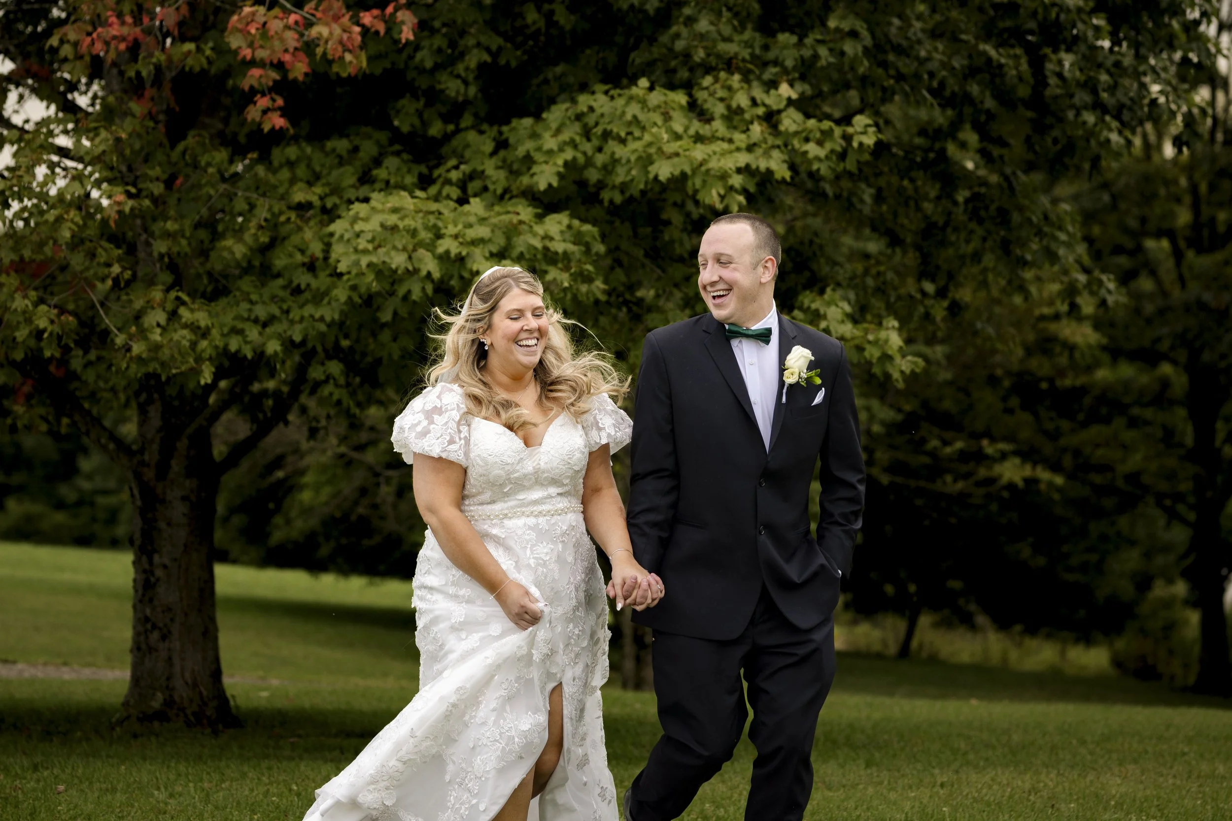 A bride and groom walking hand in hand outdoors, smiling and laughing, with trees in the background.