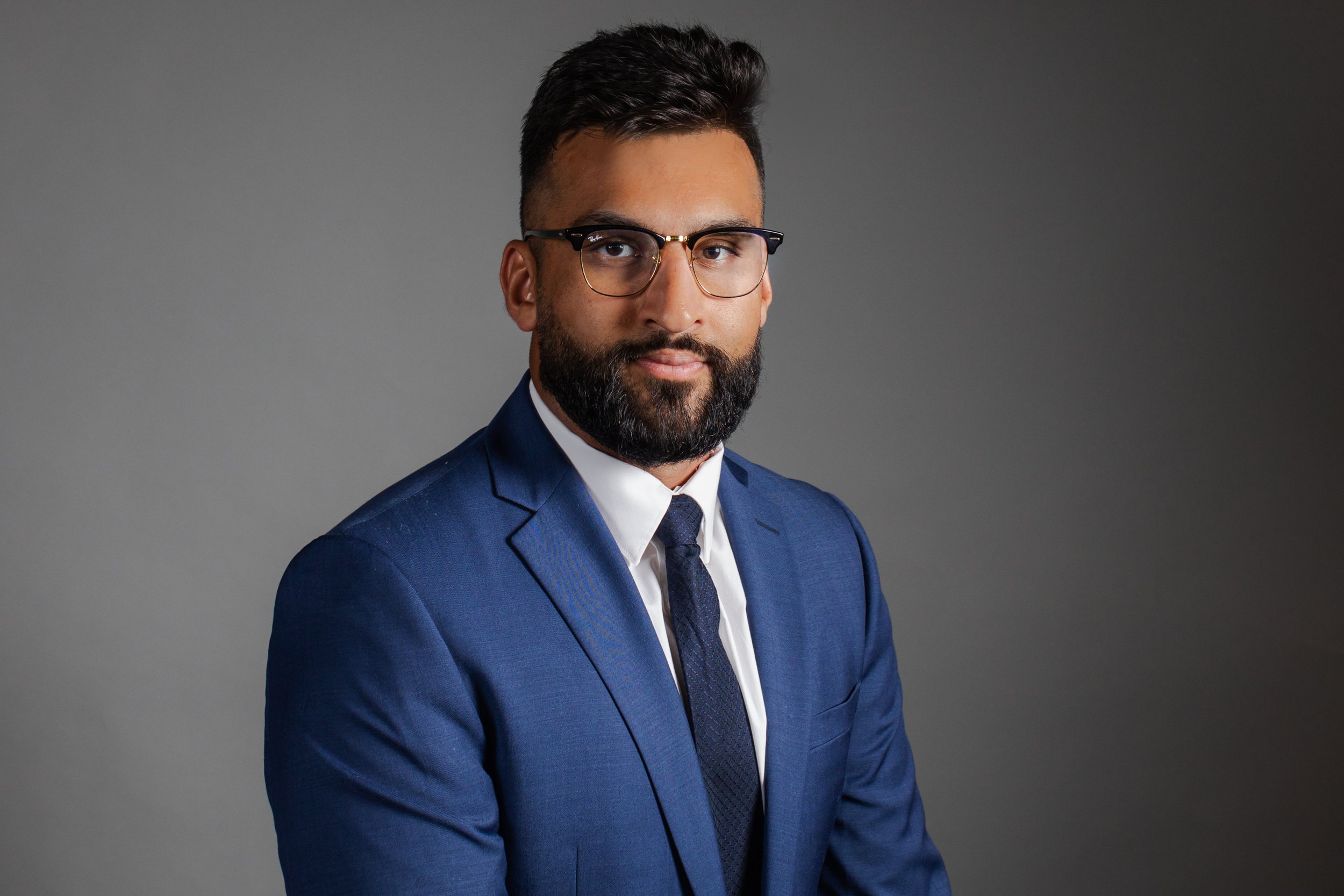 Professional portrait of a man wearing a blue suit, white shirt, and dark tie, with glasses and a beard, against a gray background.