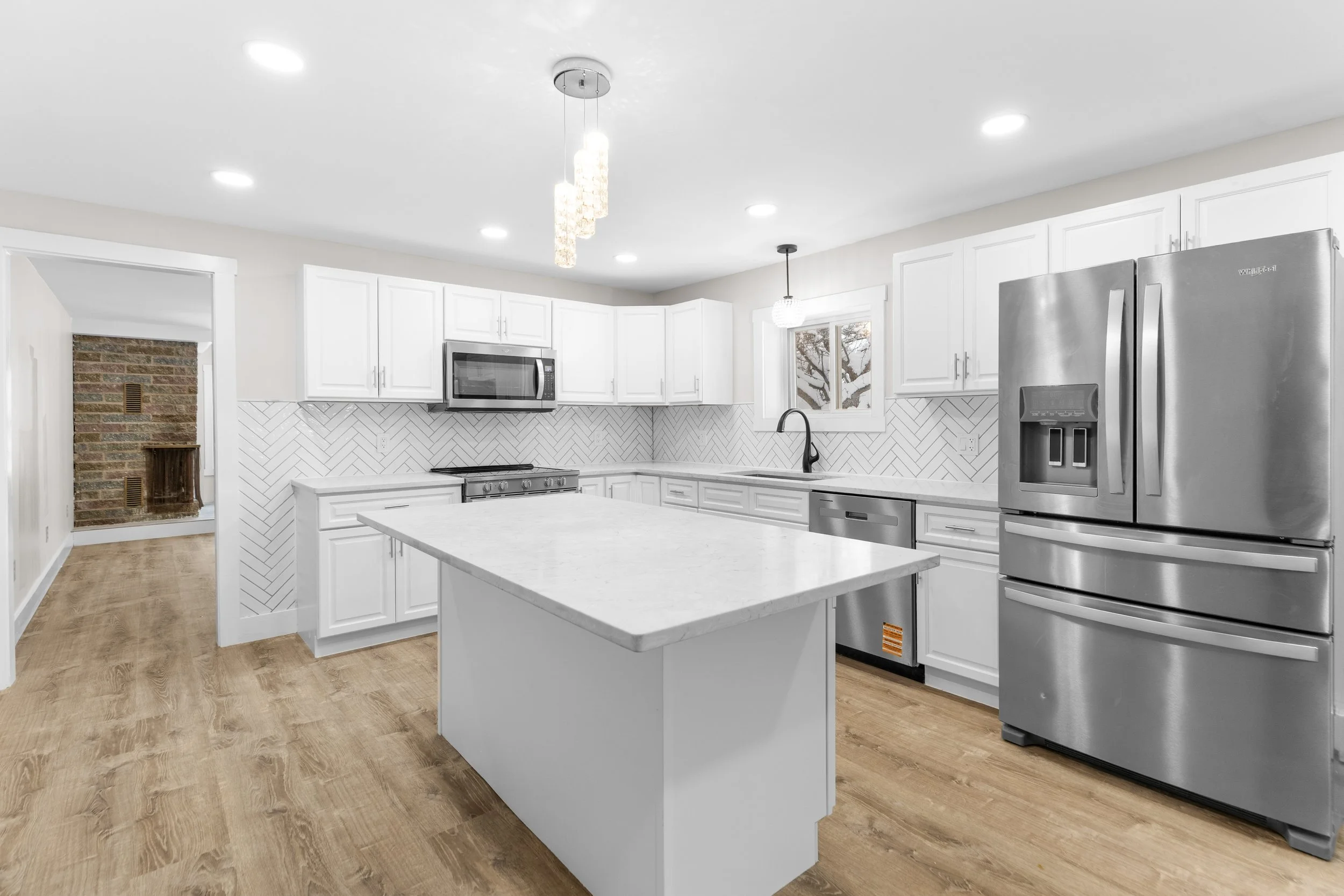 Modern kitchen with white cabinets, stainless steel appliances, light wood flooring, an island with a white countertop, and a backsplash with white herringbone tile pattern.