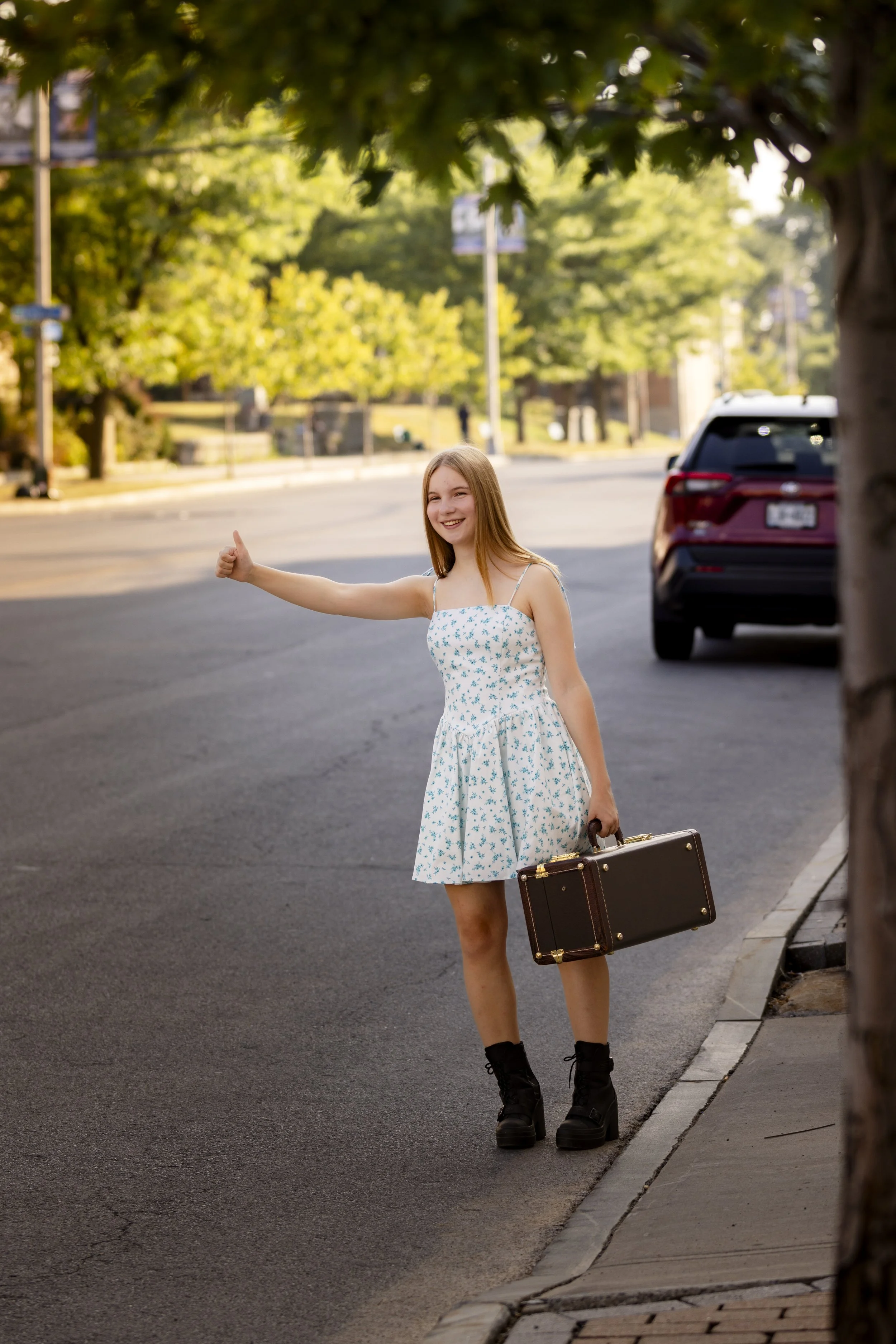 A young woman standing on a street curb, wearing a white dress with blue floral pattern, black boots, and holding a vintage briefcase, waving and giving a thumbs-up with a smile.