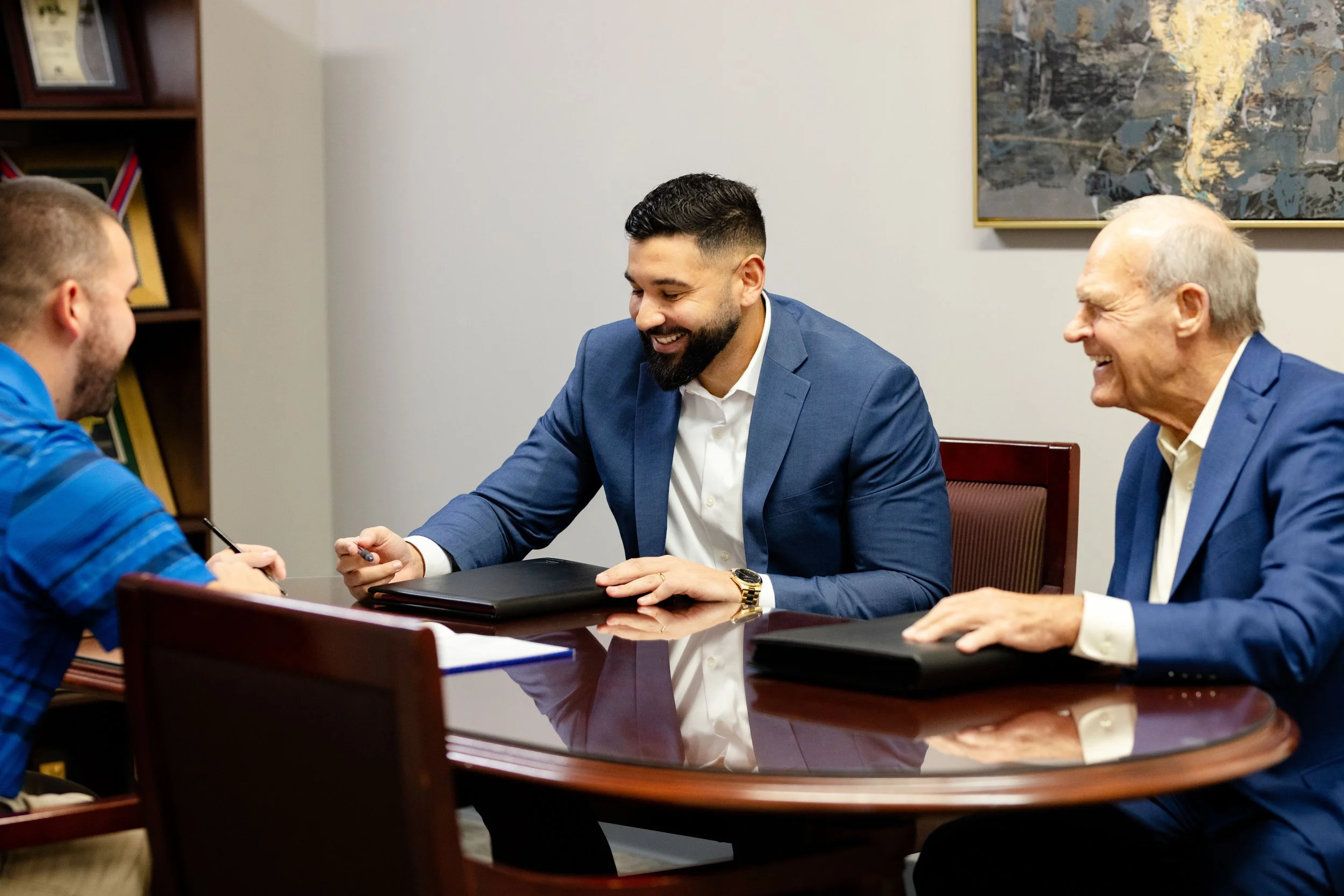 Three men in business suits sitting at a conference table, engaged in a discussion and smiling, with two of them holding pens and a closed black folder in front of each of them.