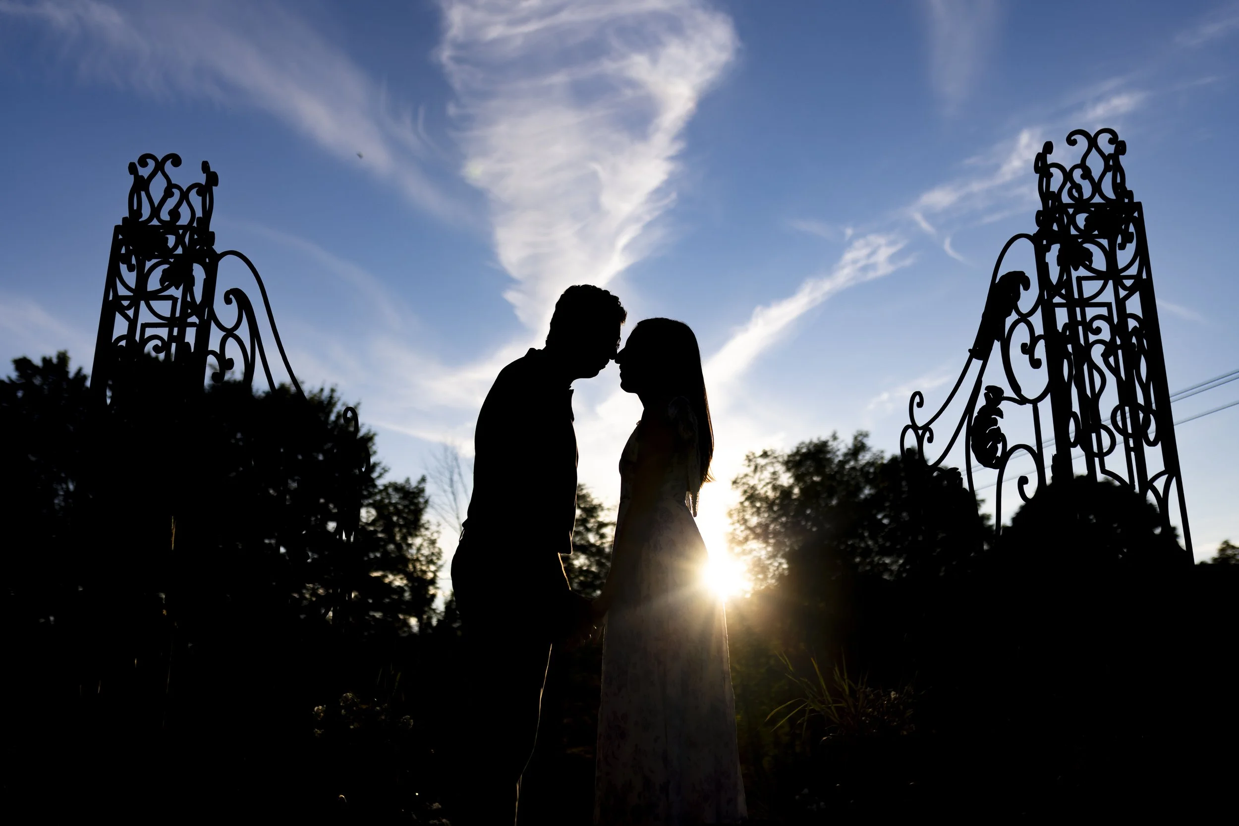 Silhouette of a couple standing face to face and holding hands outdoors during sunset, with ornate metal gates on either side and the sun low on the horizon behind them.