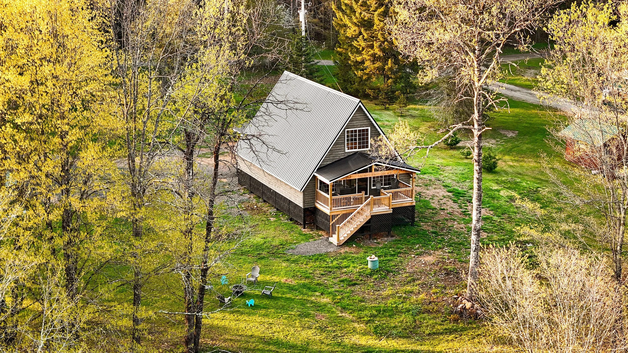 A cozy wooden house with a metal roof and a front porch situated in a lush, green yard with trees and outdoor furniture.