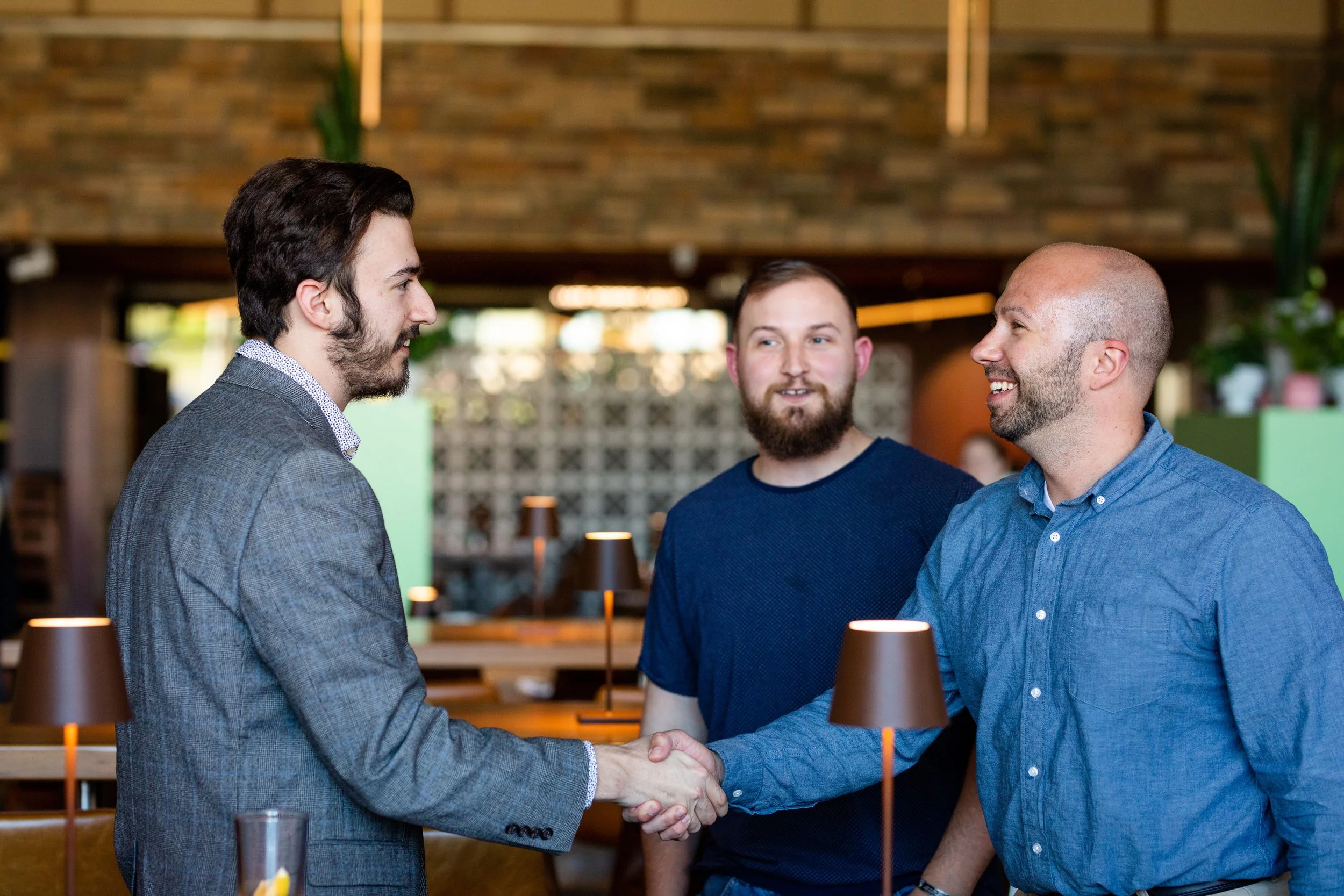 Three men in a restaurant, two of them shaking hands and smiling, while the third observes. They are casually dressed, and the restaurant has warm lighting and modern decor.