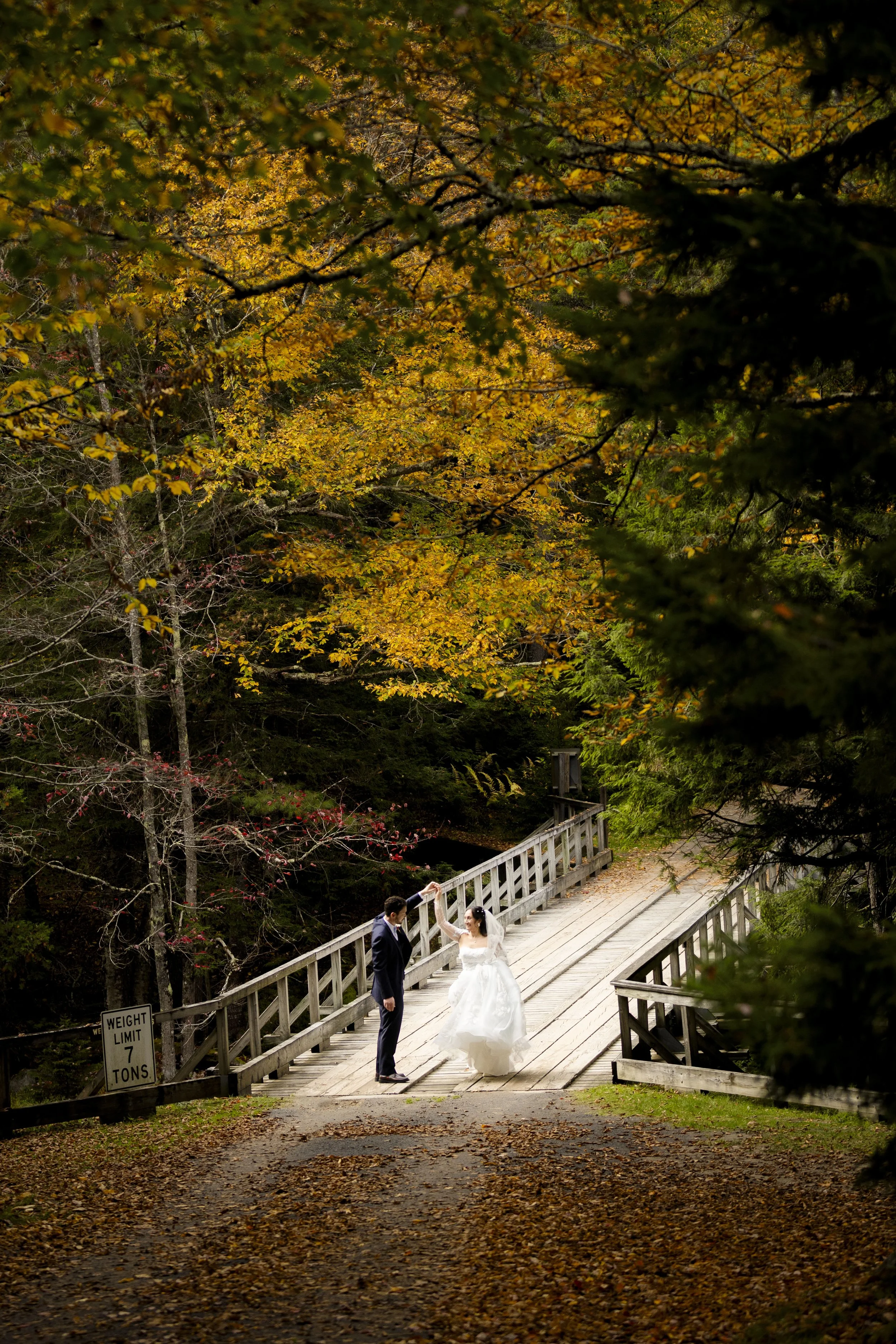 A bride and groom dancing on a wooden bridge surrounded by autumn trees, with fallen leaves on the ground.