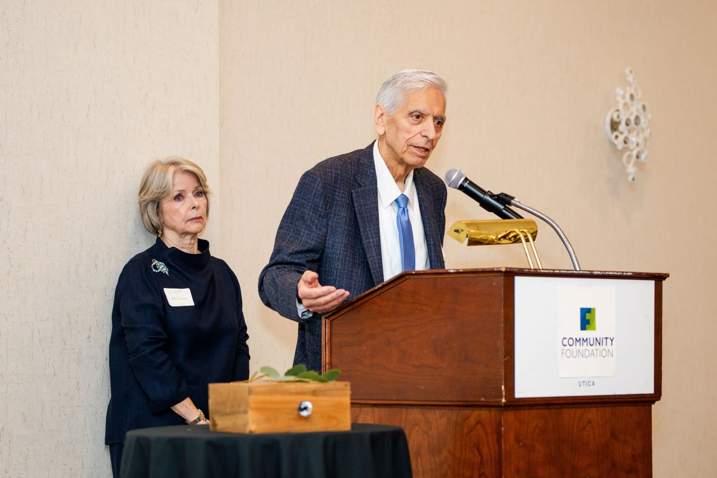An elderly man in a gray suit and blue tie speaking at a podium with a microphone, a woman with blonde hair in a dark outfit standing beside him, and a sign on the podium that reads 'Community Foundation Utica'.