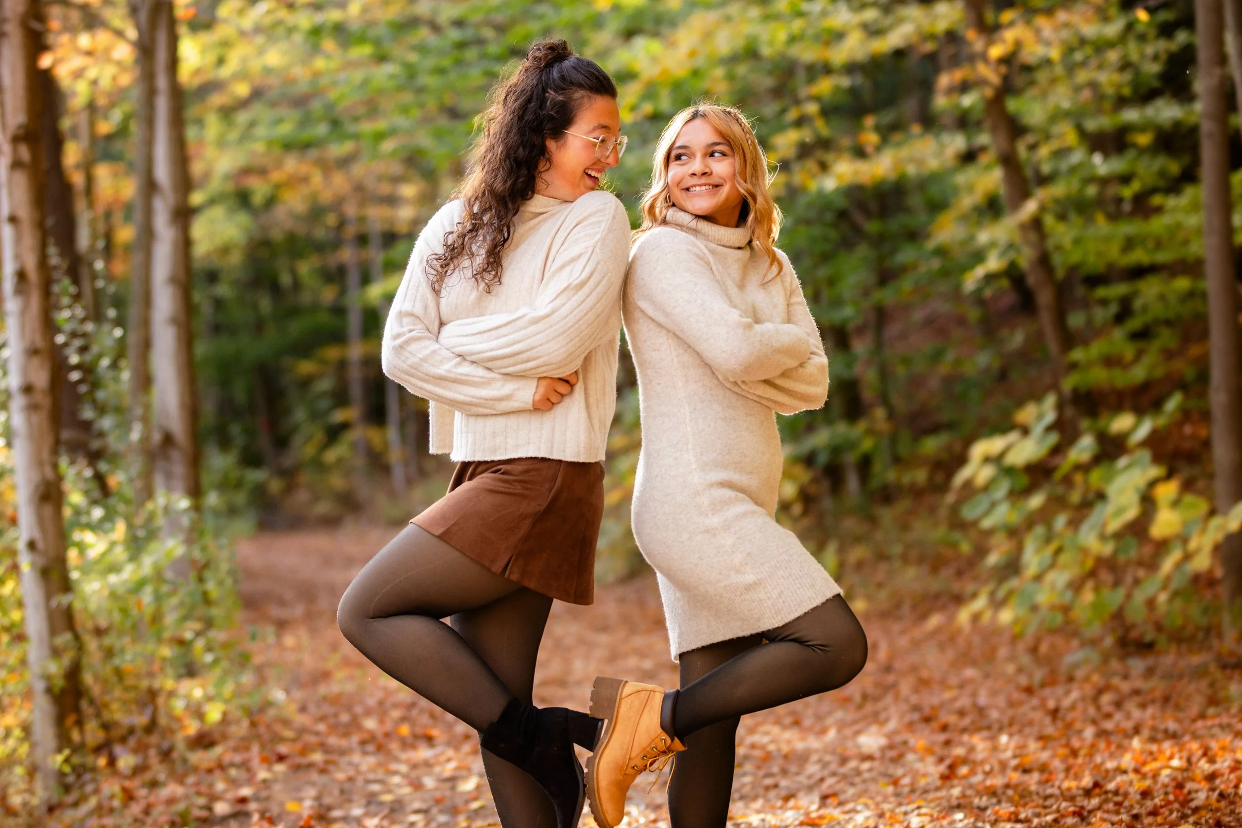 Two women wearing cozy sweaters and leggings enjoying fall walk in a leaf-covered forest with trees and autumn foliage.