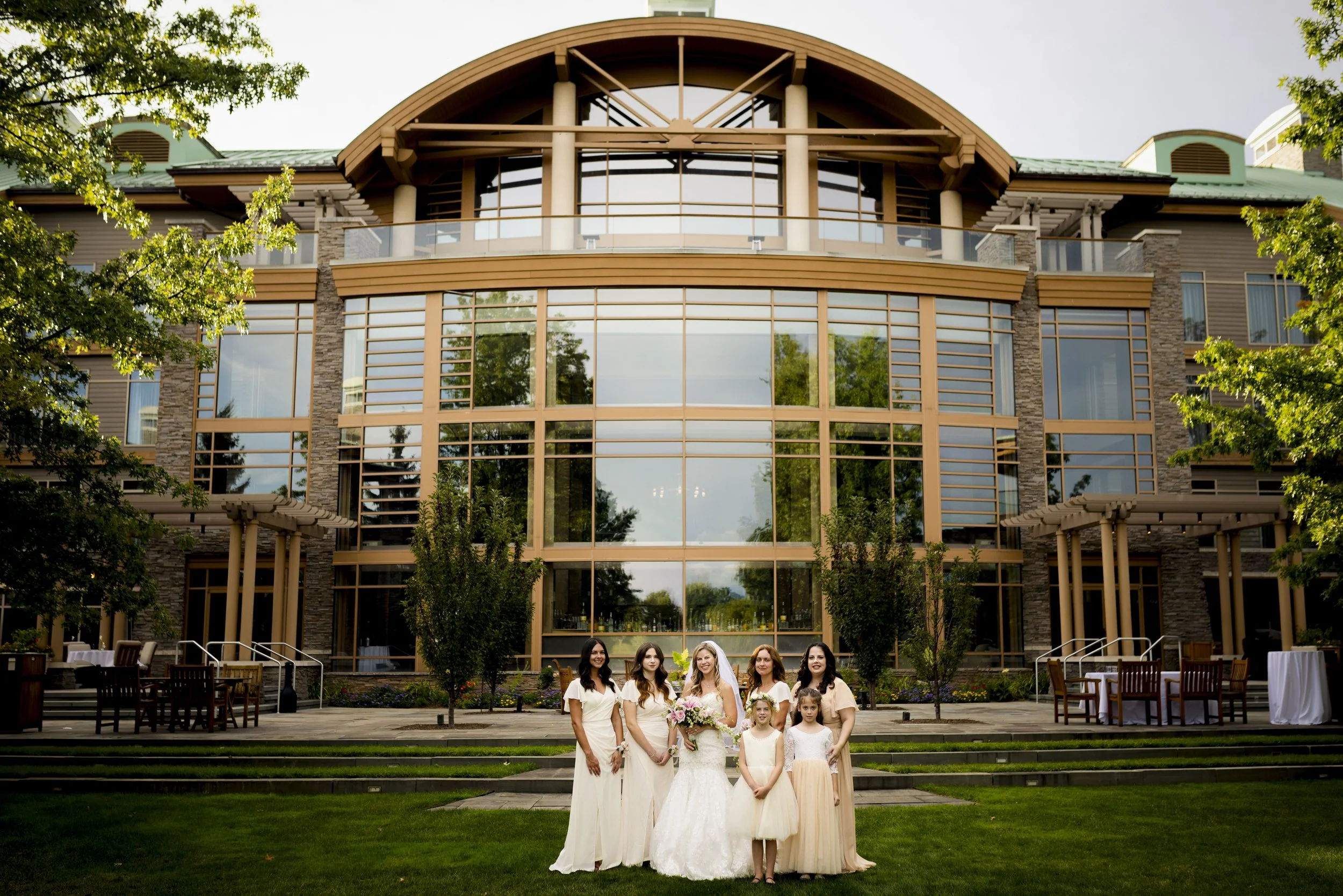 Group of six women, including a bride in a white wedding dress holding a bouquet, standing on grass in front of a modern glass building with trees and outdoor furniture.