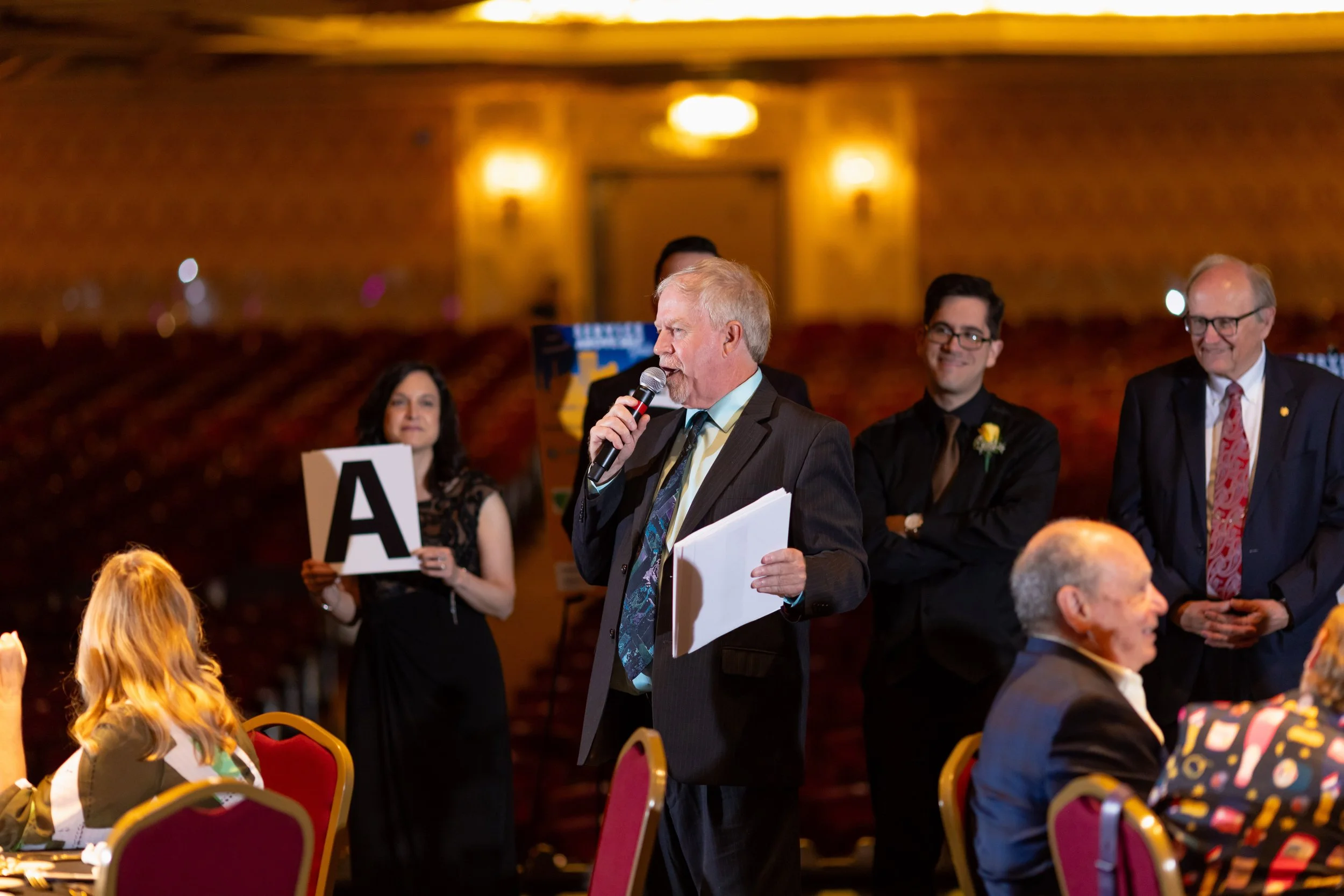 An older man with white hair and a beard speaking into a microphone while holding a stack of papers at an indoor event. Behind him are three people standing, including a woman holding a sign with the letter 'A', and two men dressed in formal attire. 