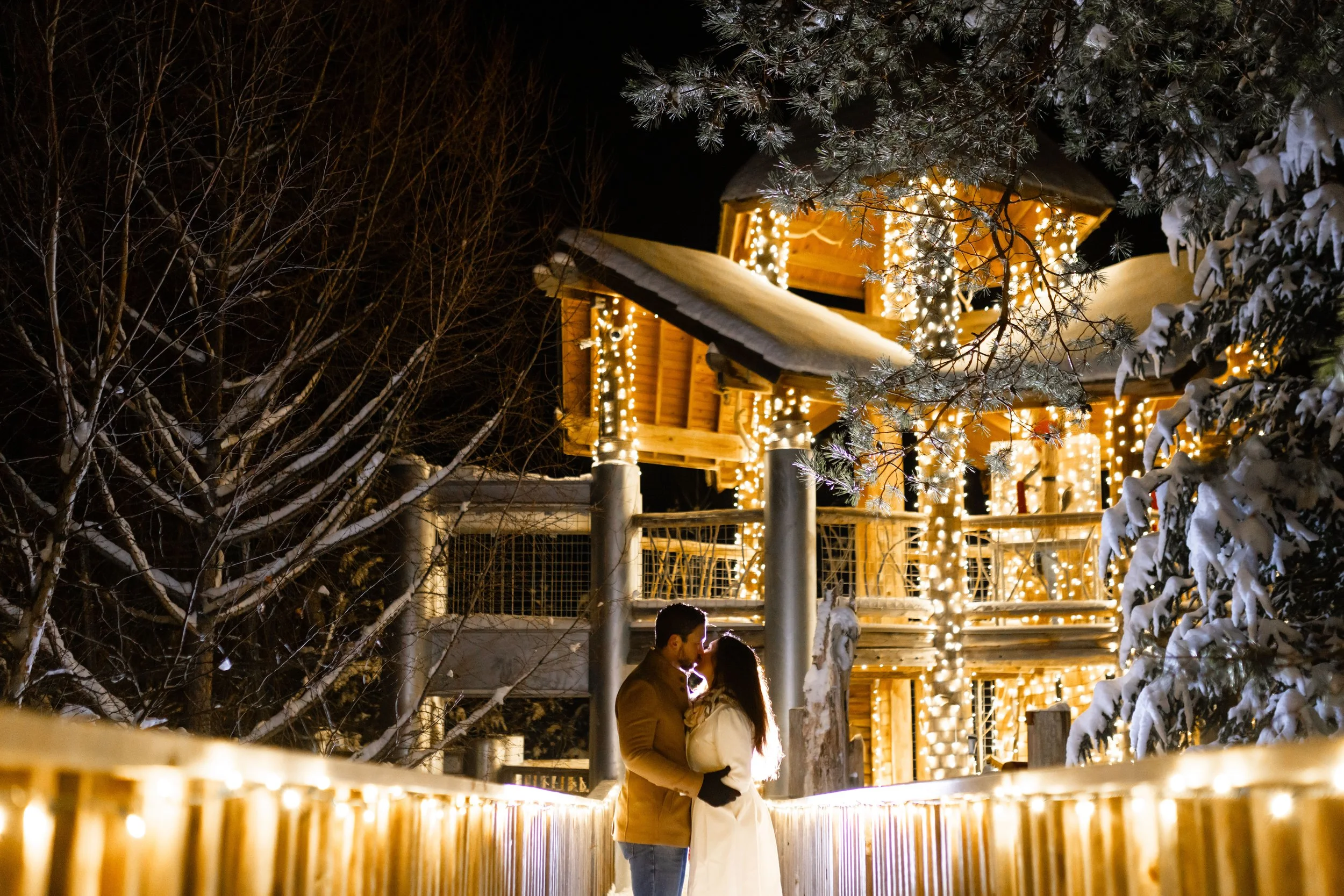 A couple is standing close and sharing a kiss on a wooden bridge decorated with warm string lights. In the background, there is a wooden house with more string lights, and snow is on the roof and surrounding trees.