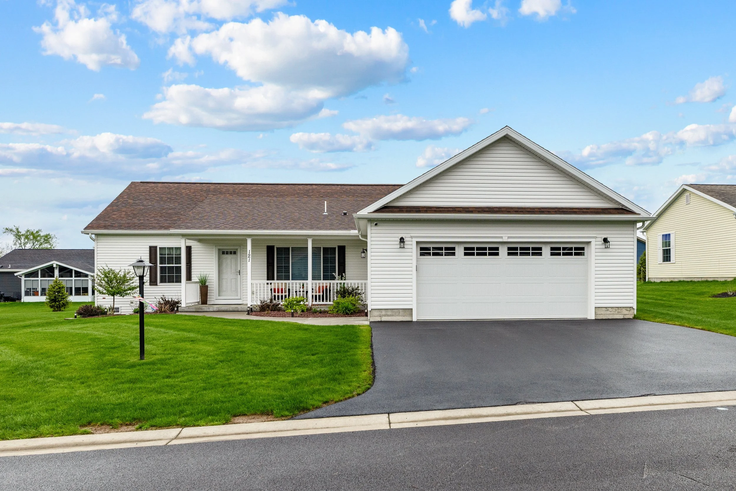 Front view of a suburban house with white siding, a brown shingle roof, a front porch with seating, and a driveway leading to an attached garage, with green lawn and a blue sky with clouds.