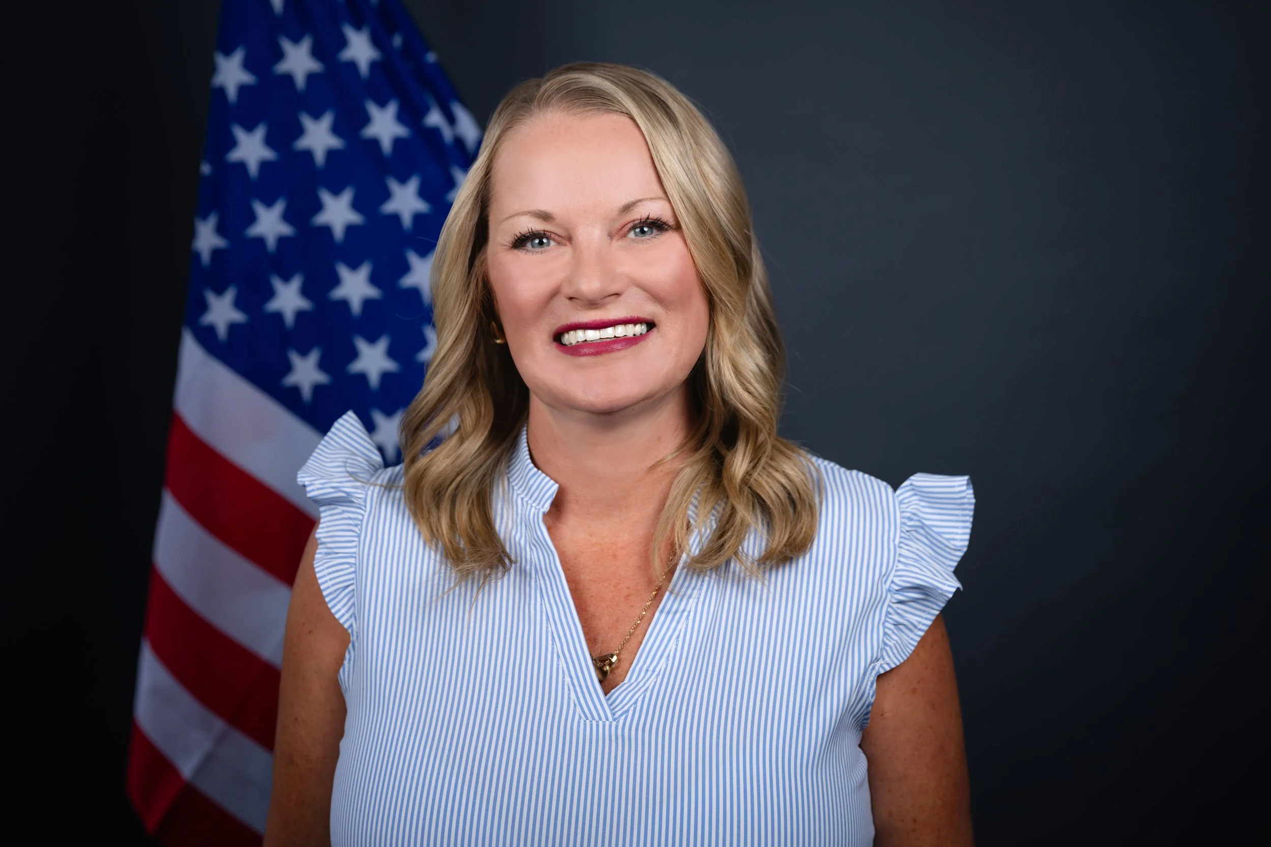 A woman with blonde hair smiling, standing in front of an American flag and a dark background, wearing a blue and white striped blouse with ruffled sleeves.