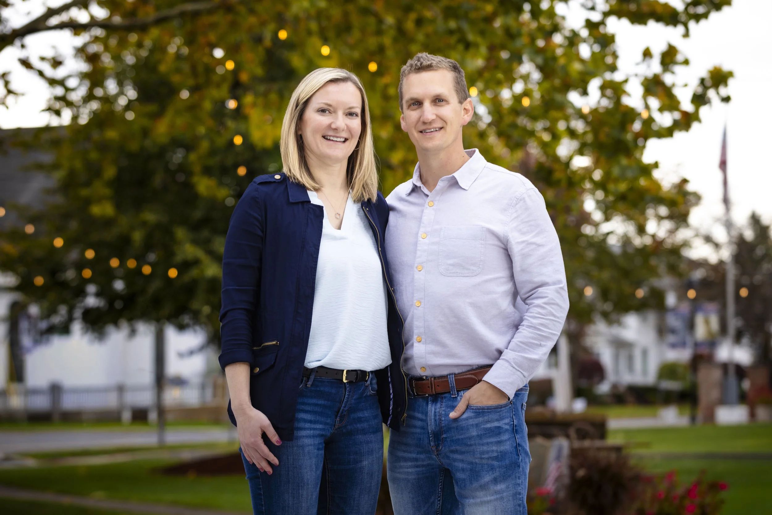 A smiling couple standing outdoors in front of trees with autumn leaves, with string lights and houses in the background.