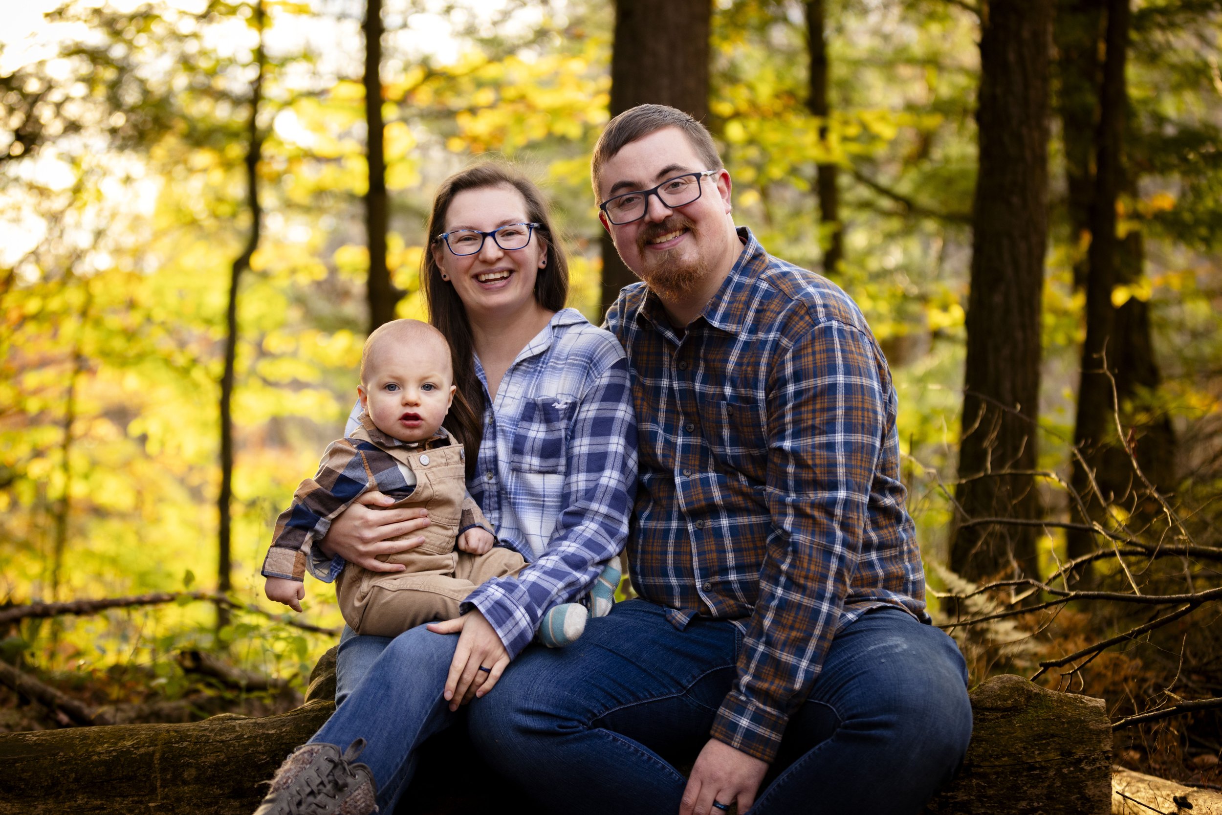 Family of three, two adults and a toddler, sitting on a log in a wooded area with autumn foliage, smiling at the camera.