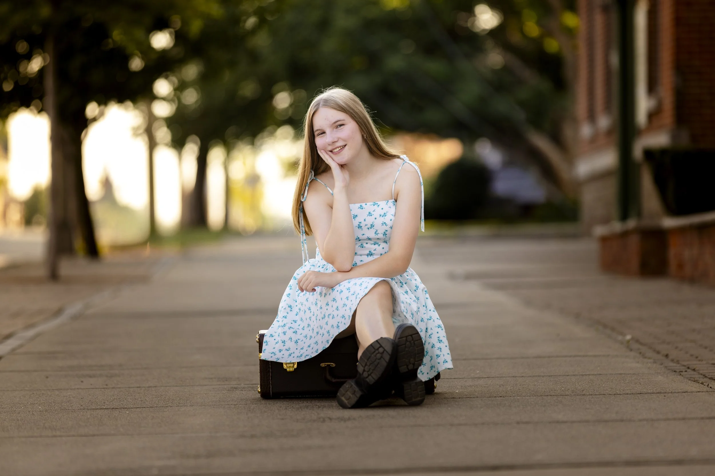 A young woman with long blonde hair in a blue floral dress sitting on a small suitcase on a sidewalk, smiling with her hand resting on her cheek. The background shows trees and a brick building, with warm lighting indicating late afternoon.