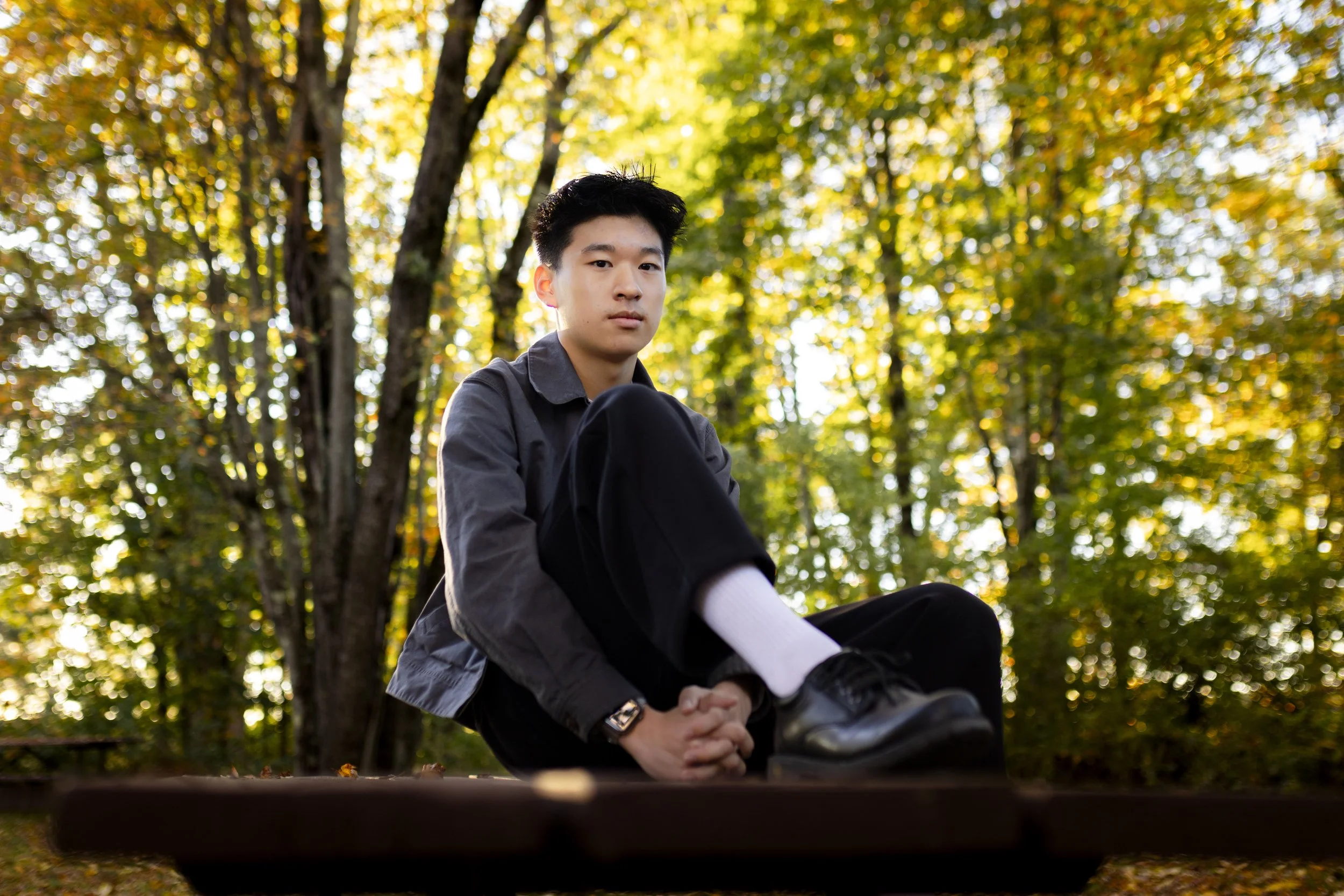 A young man sitting on a bench outdoors in a park with autumn trees in the background.