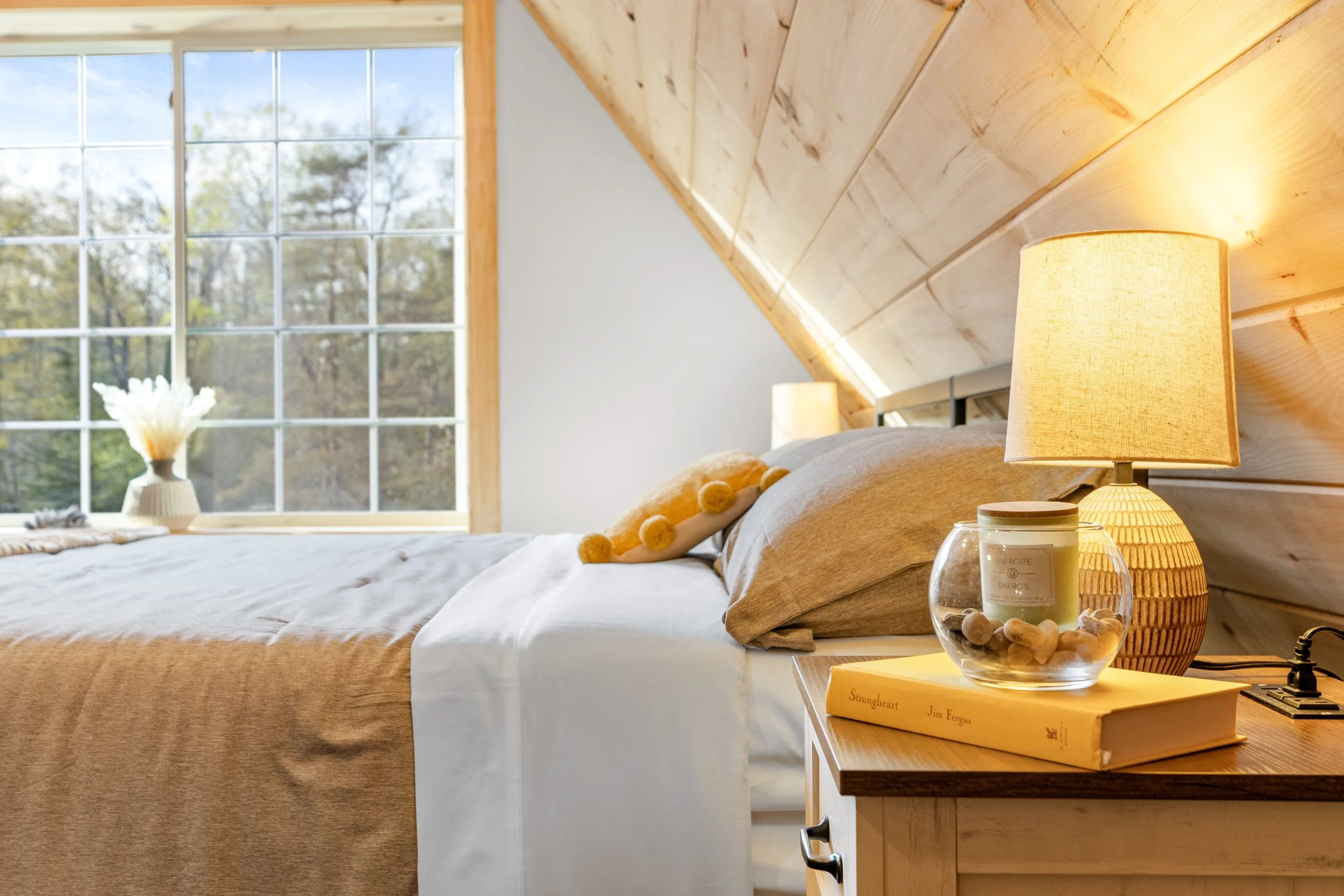 A cozy bedroom with a large window, beige bedspread, and a nightstand with a lamp, candle, and book, under a sloped wooden ceiling.