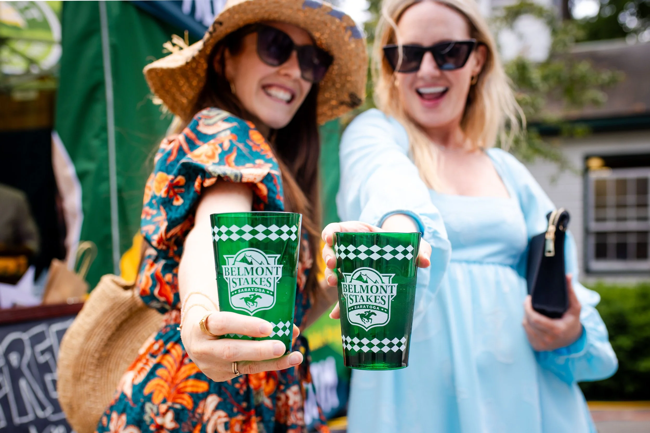 Two women smiling and holding green glasses with 'Belmont Stakes Saratoga' logo at an outdoor event.
