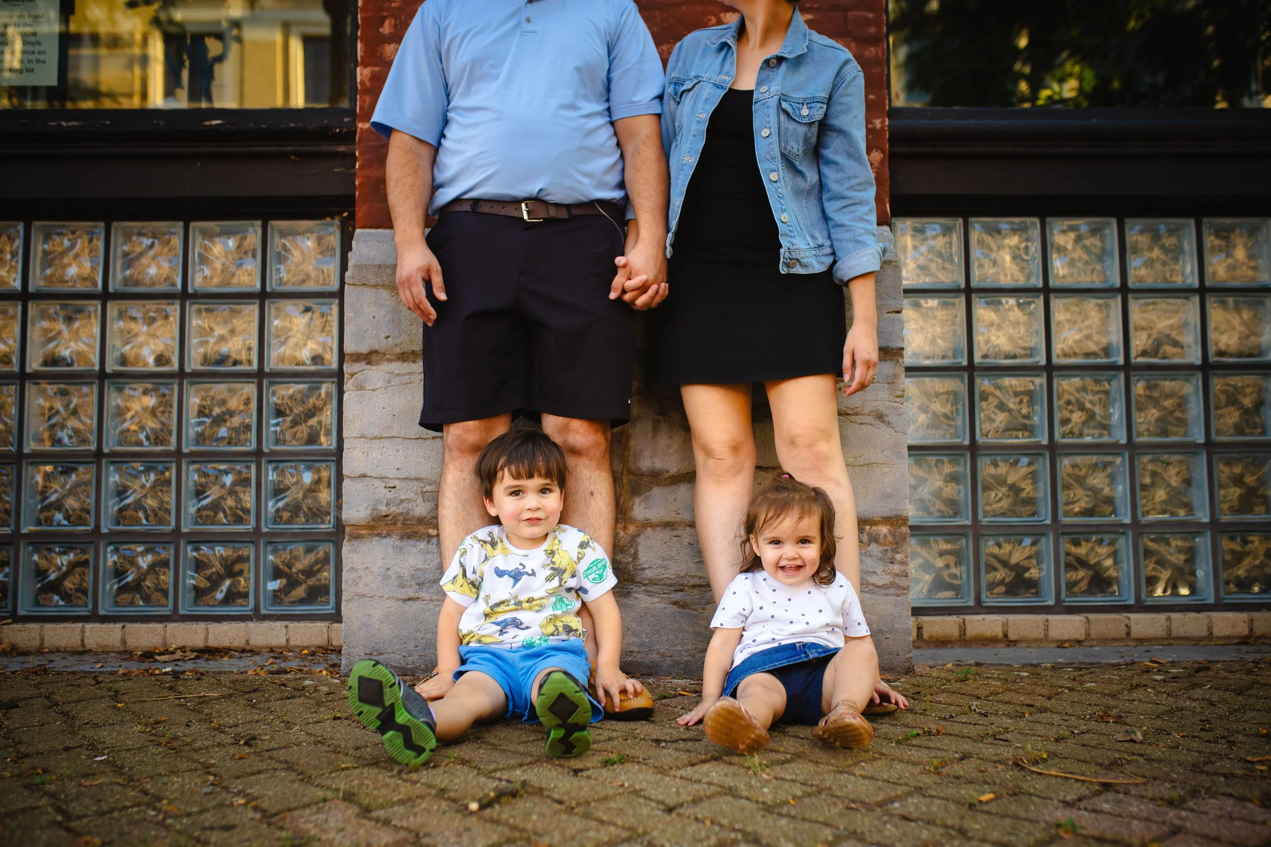 A family of four standing outside on a brick sidewalk in front of a building with glass block windows. The father and mother are holding hands, with the children sitting on the ground in front of them. The father is wearing a blue shirt and black sho