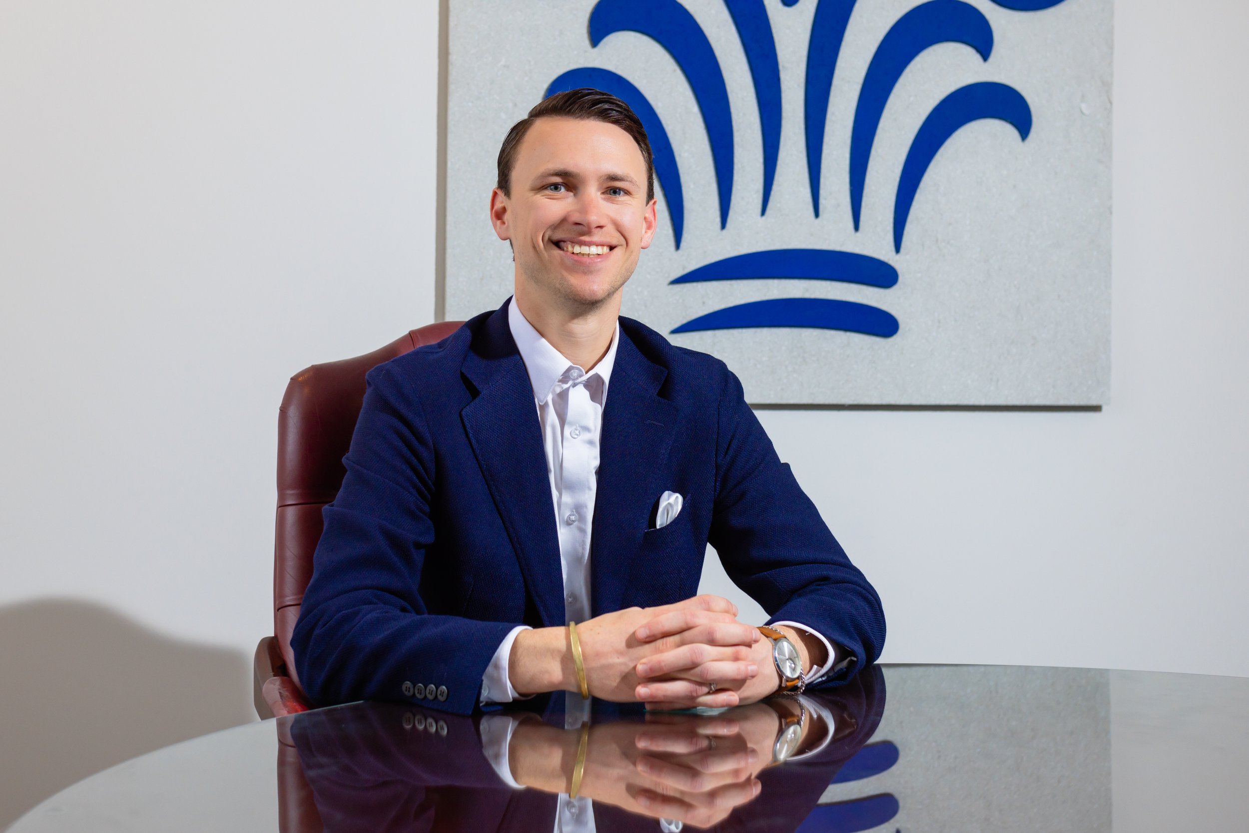 A young man with brown hair, in a navy blue blazer and white shirt, sits at a dark reflective conference table in an office, smiling at the camera. Behind him is a wall with a large blue abstract logo.