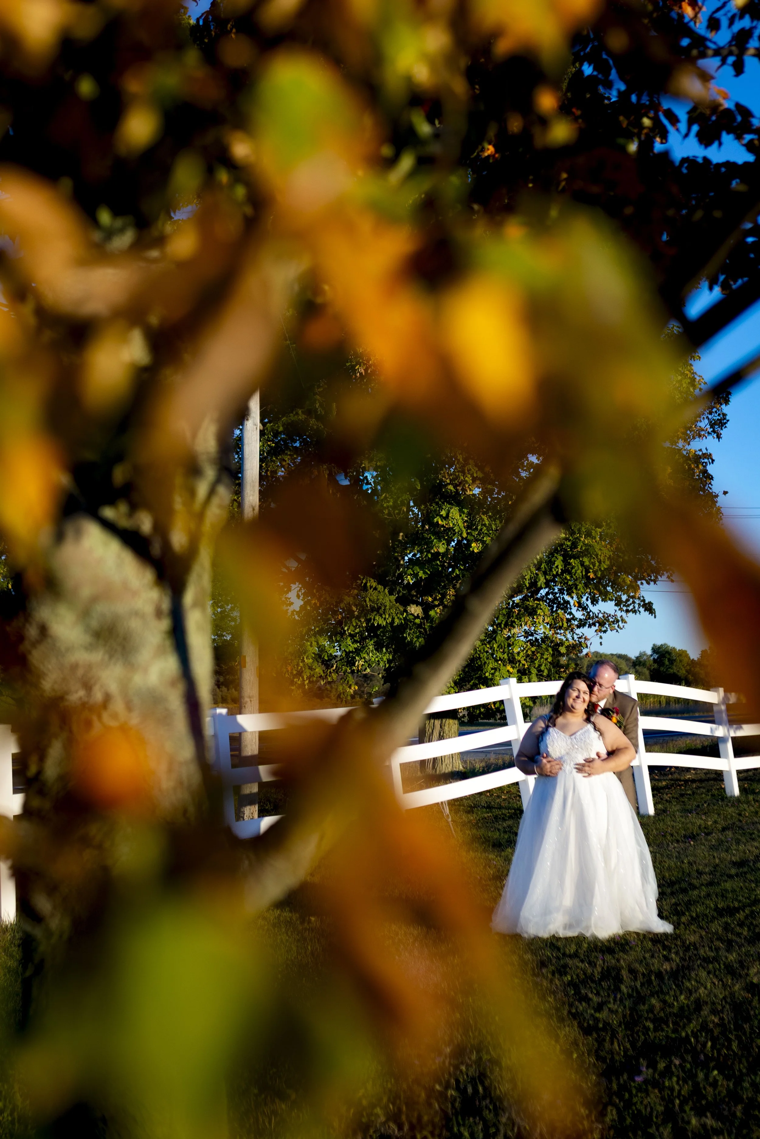 A bride and groom standing together outdoors in front of a white fence, with autumn leaves in the foreground and a clear blue sky in the background.