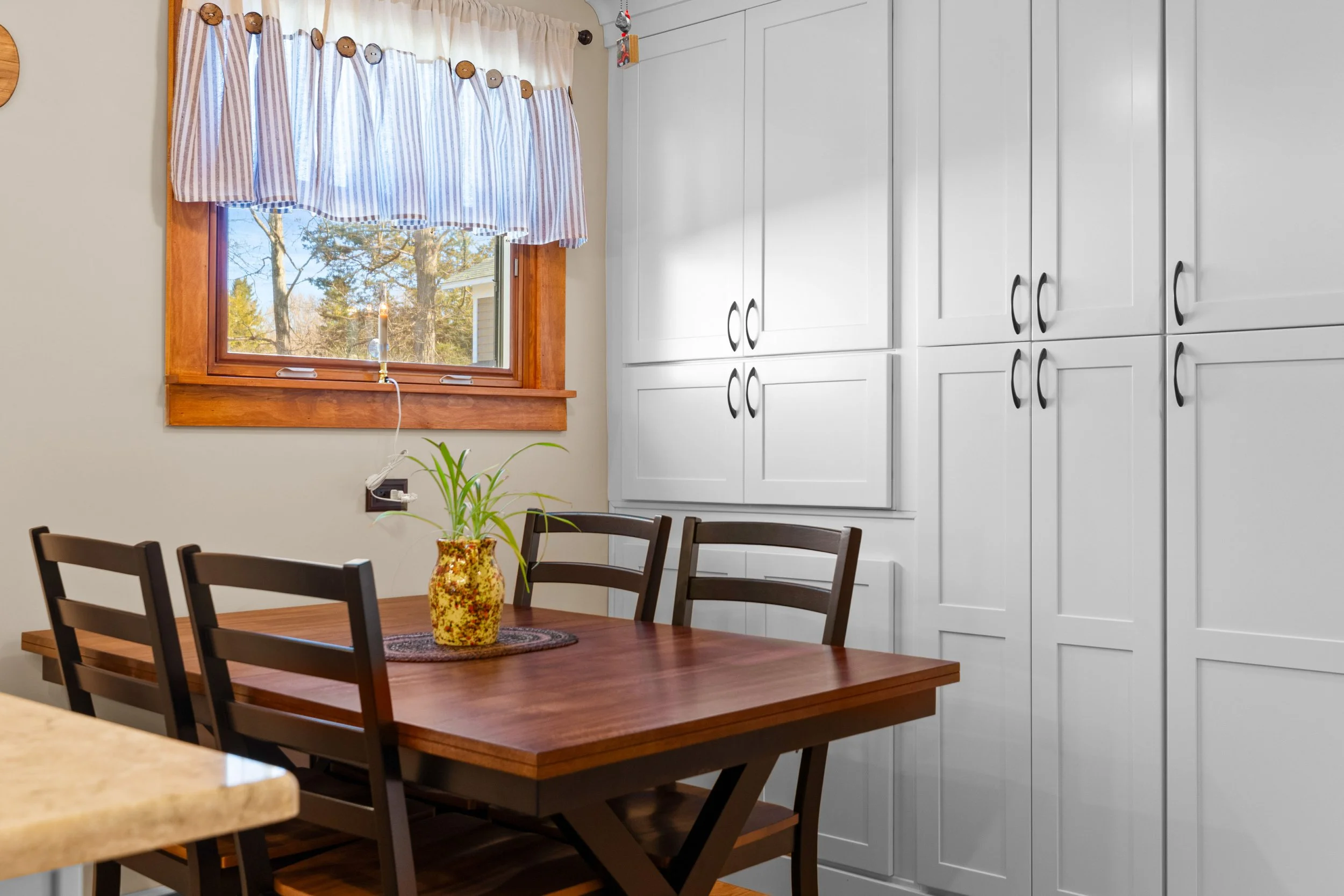 A kitchen dining area with a wooden table, four dark wood chairs, a yellow patterned vase with green plants, and a window with striped curtains overlooking trees outside.