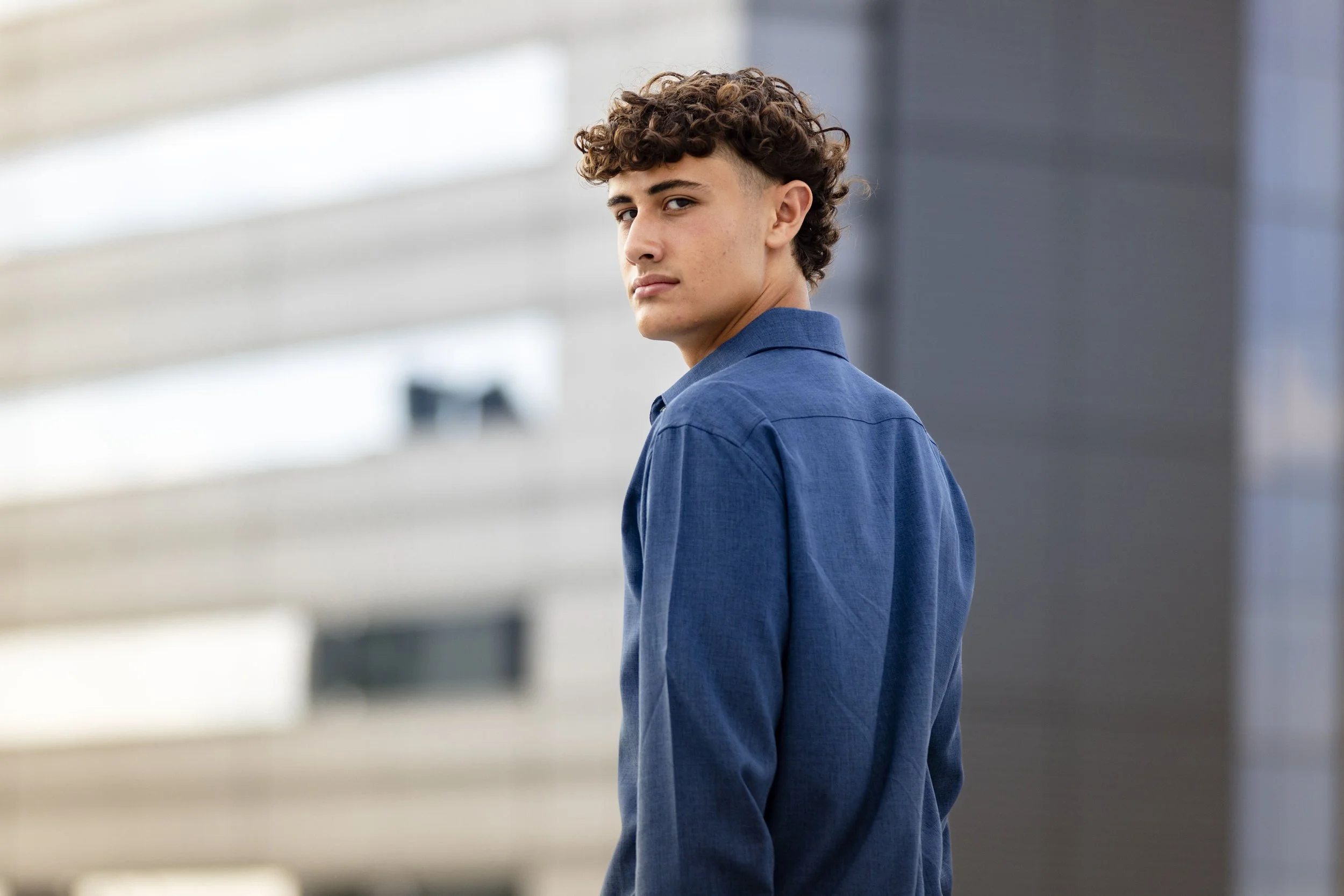 A young man with curly brown hair, wearing a blue button-up shirt, stands outdoors with a modern building in the background, looking over his shoulder at the camera.