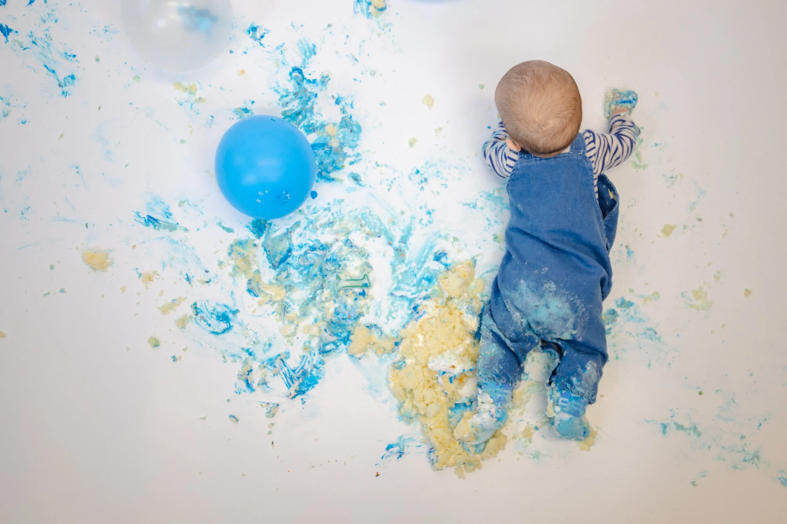 Child in blue overalls and striped shirt lying on white floor covered in blue and yellow cake or frosting, with blue and clear balloons nearby.