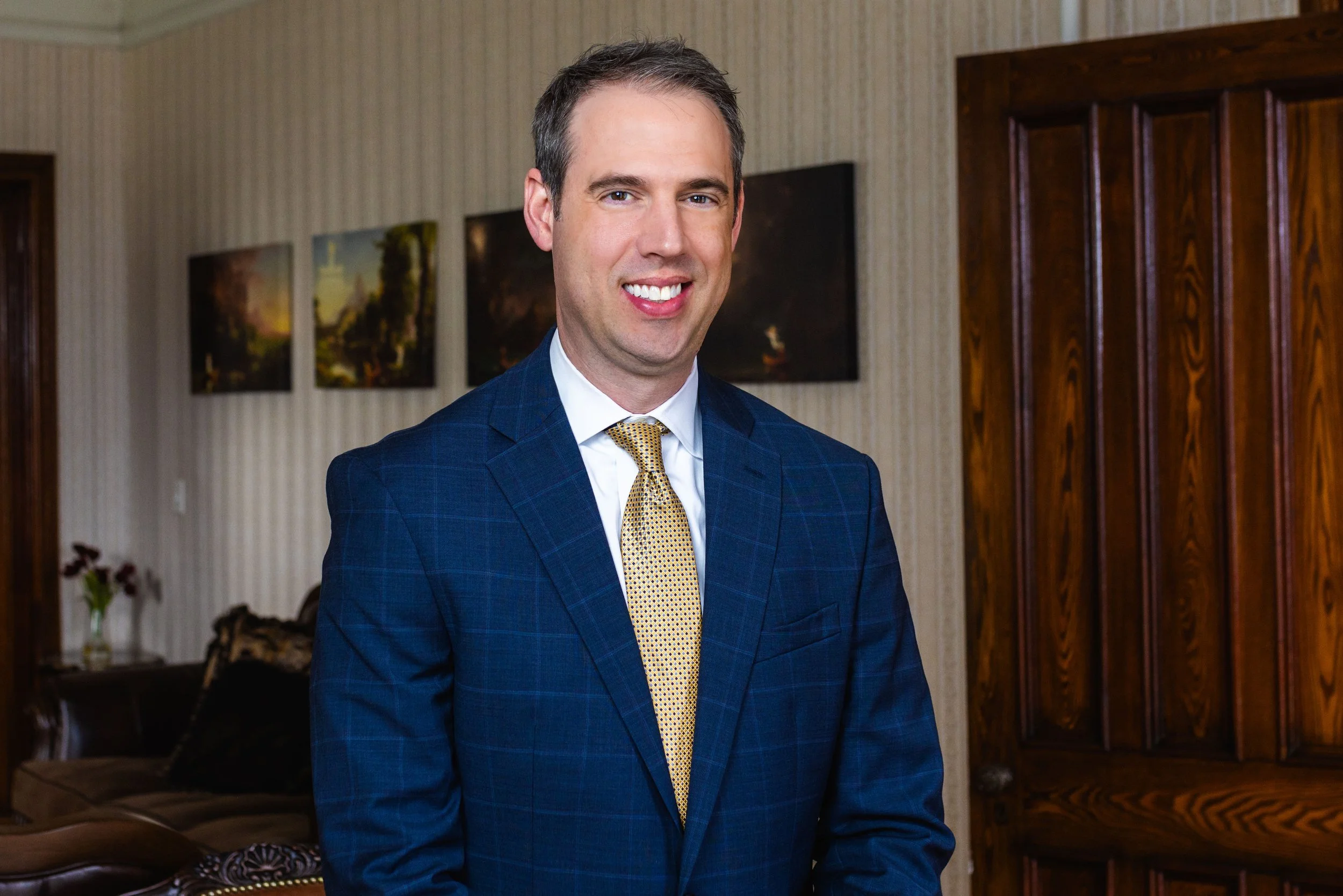 A man in a blue checkered suit with a white shirt and a gold tie, smiling in a room with framed landscape pictures on the wall and wooden furniture.