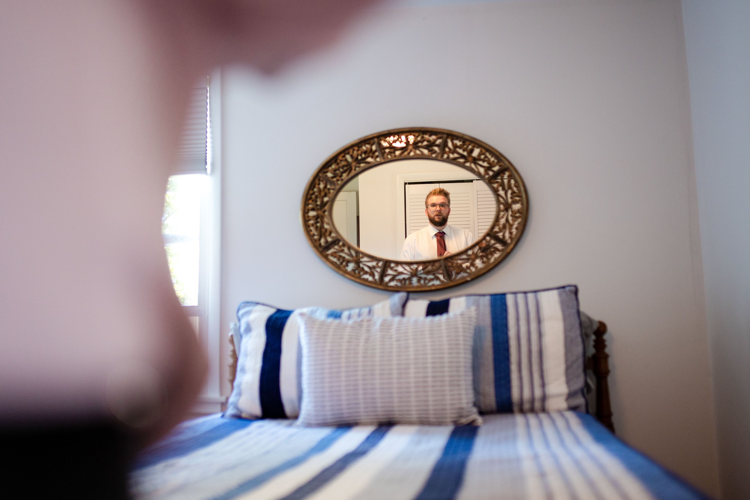 A man with glasses and a beard, wearing a white shirt and red tie, is seen in a mirror hanging above a bed with striped blue and white bedding.
