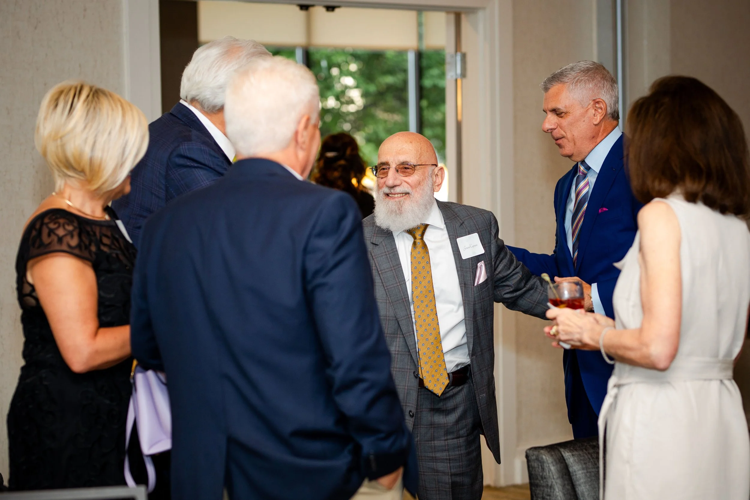 Group of professionally dressed people engaging in conversation at a social or business event inside a room with an open door leading outside to greenery.